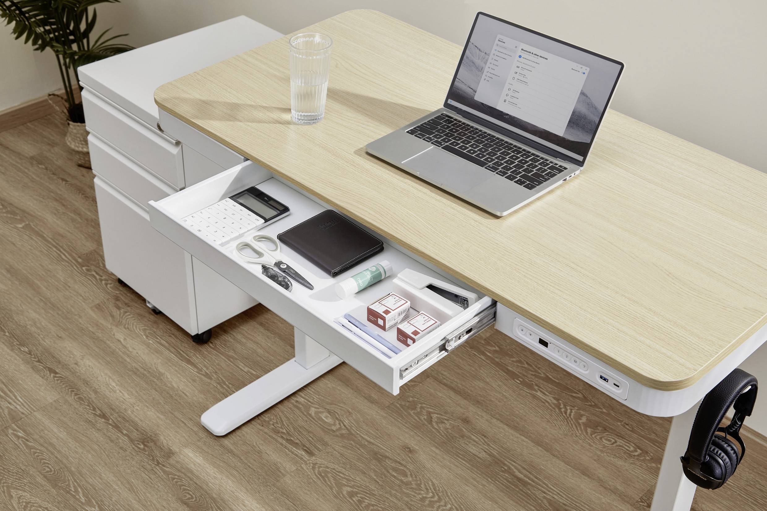 Een moderne bureau met een laptop en een glas water erop. Een opengetrokken lade laat kantoorbenodigdheden zien.