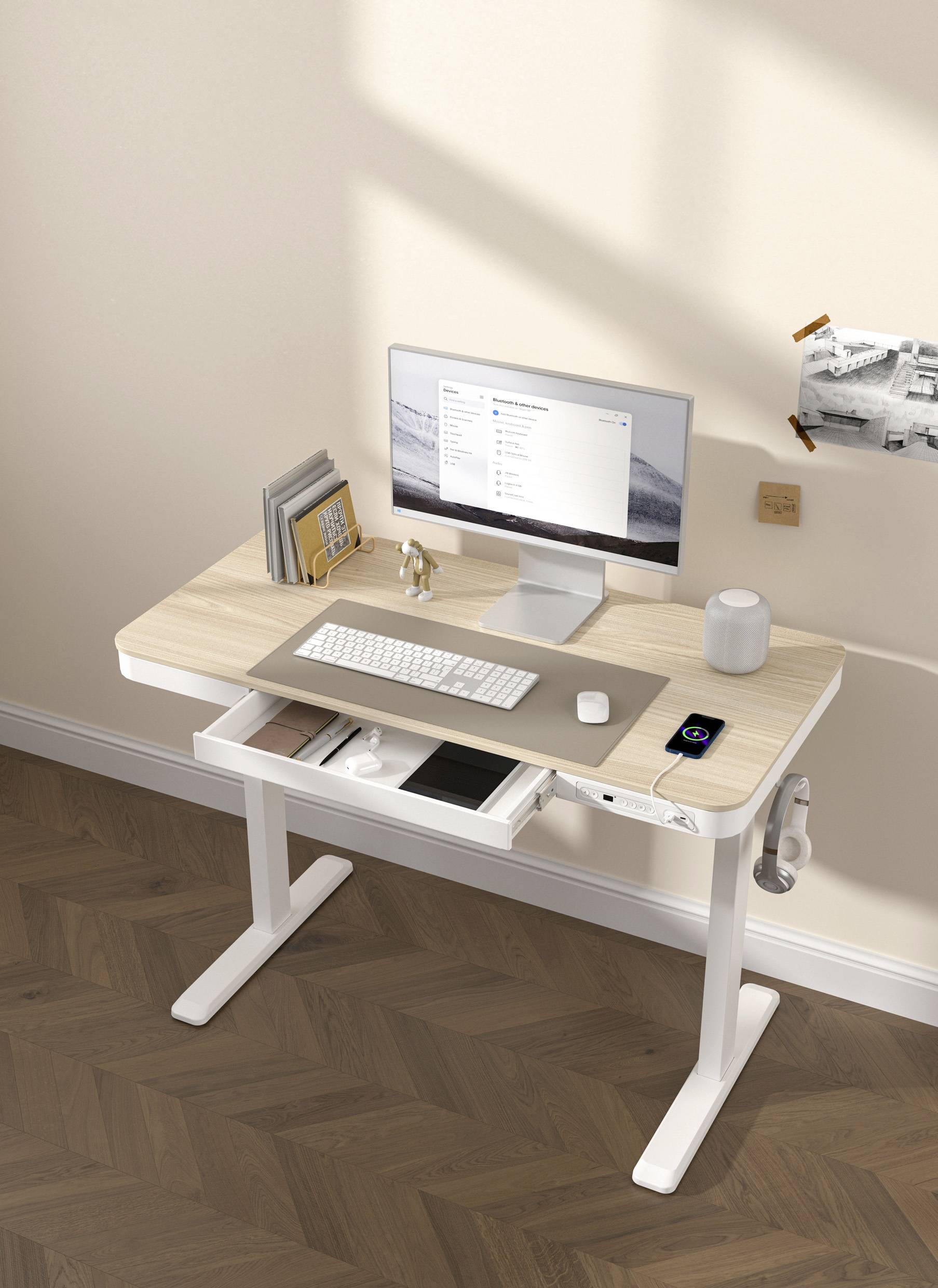 Een moderne werkplek met monitor, toetsenbord, muis en smartphone. Aan de linkerkant staan boeken en rechts hangt een schilderij aan de muur.