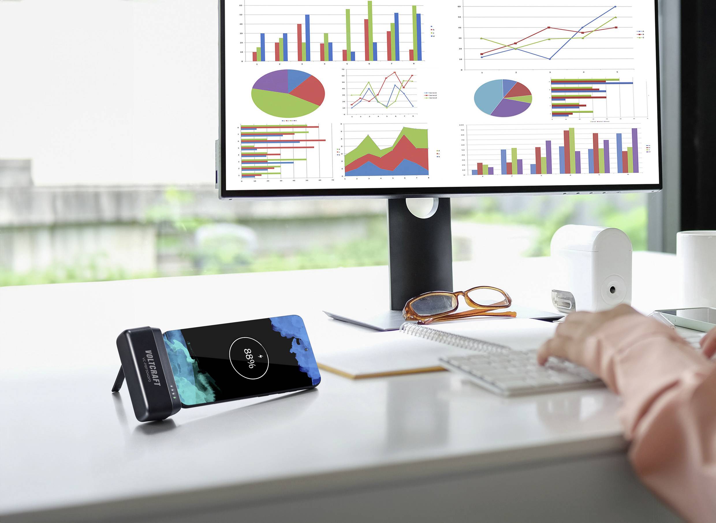 Een persoon werkt aan een bureau met grafieken op een computerscherm. Een smartphone wordt draadloos opgeladen op een standaard.
