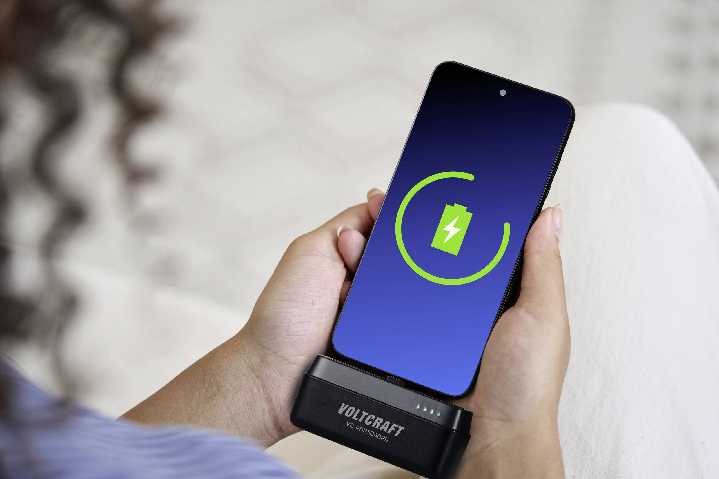 Persoon houdt smartphone met laadstatus op het scherm. Het apparaat wordt opgeladen met een VOLTCRAFT-oplader.