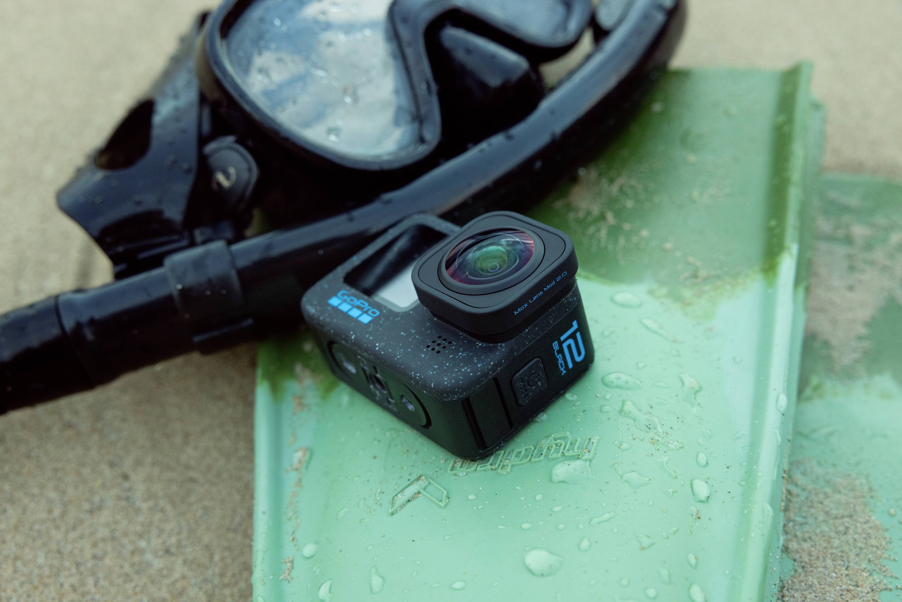 Een waterdichte actiecamera ligt op een groene zwemvin naast een duikbril op het strand, omringd door waterdruppels.