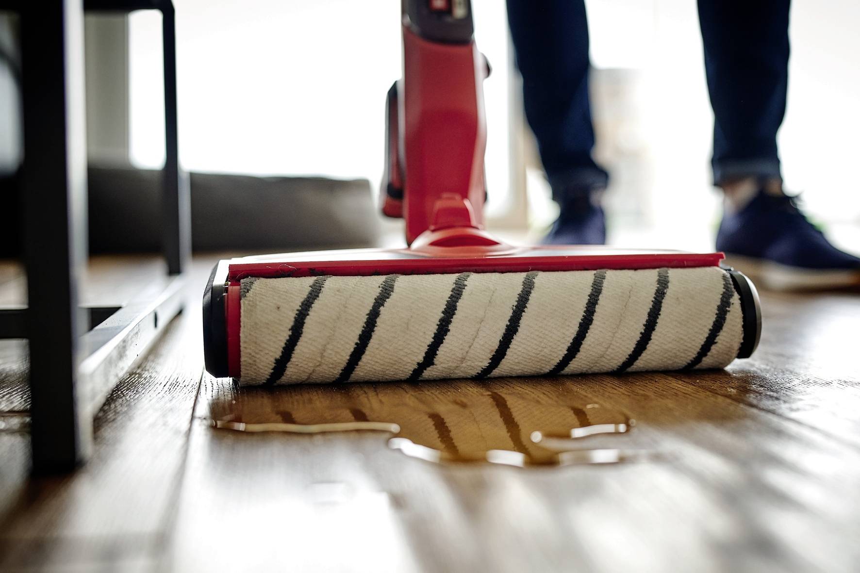 Een dweil veegt een houten vloer, terwijl water over de vloer verspreid is. Op de achtergrond zijn de onscherpe benen van een persoon te zien.