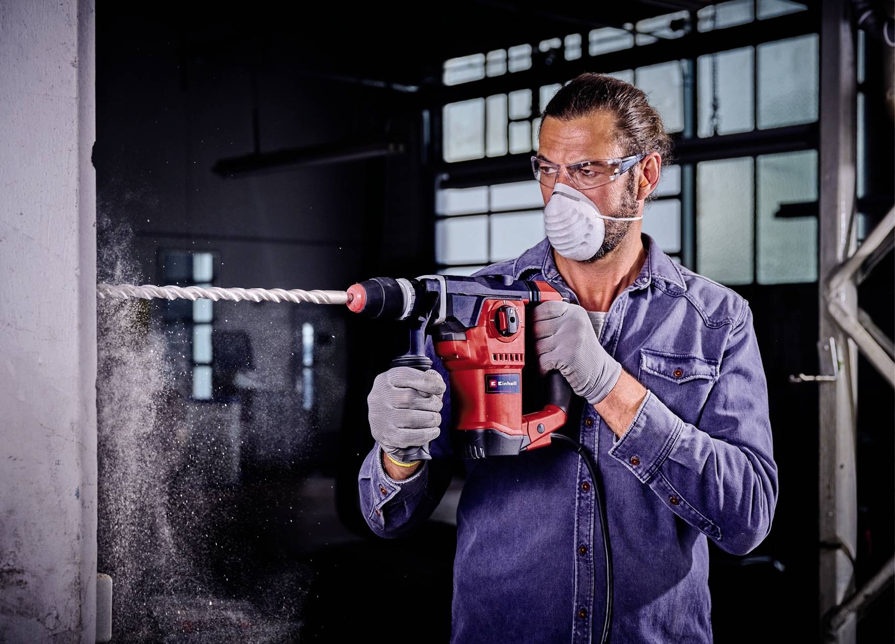 Man in blauwe spijkerjack en beschermmasker boort met een rode boormachine in een betonnen muur, stof spat rond.