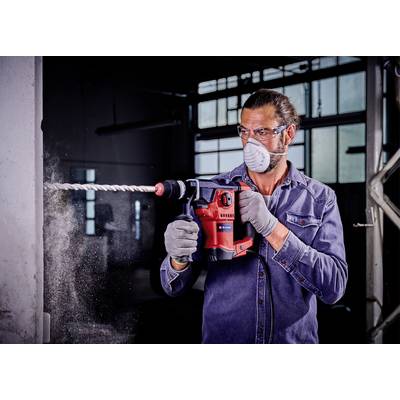 Man in blauwe spijkerjack en beschermmasker boort met een rode boormachine in een betonnen muur, stof spat rond.