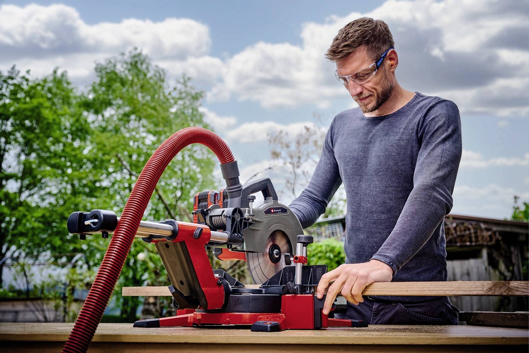Een man met veiligheidsbril werkt buiten aan een rood-zwarte afkortzaag, omringd door groene bomen en een bewolkte hemel.