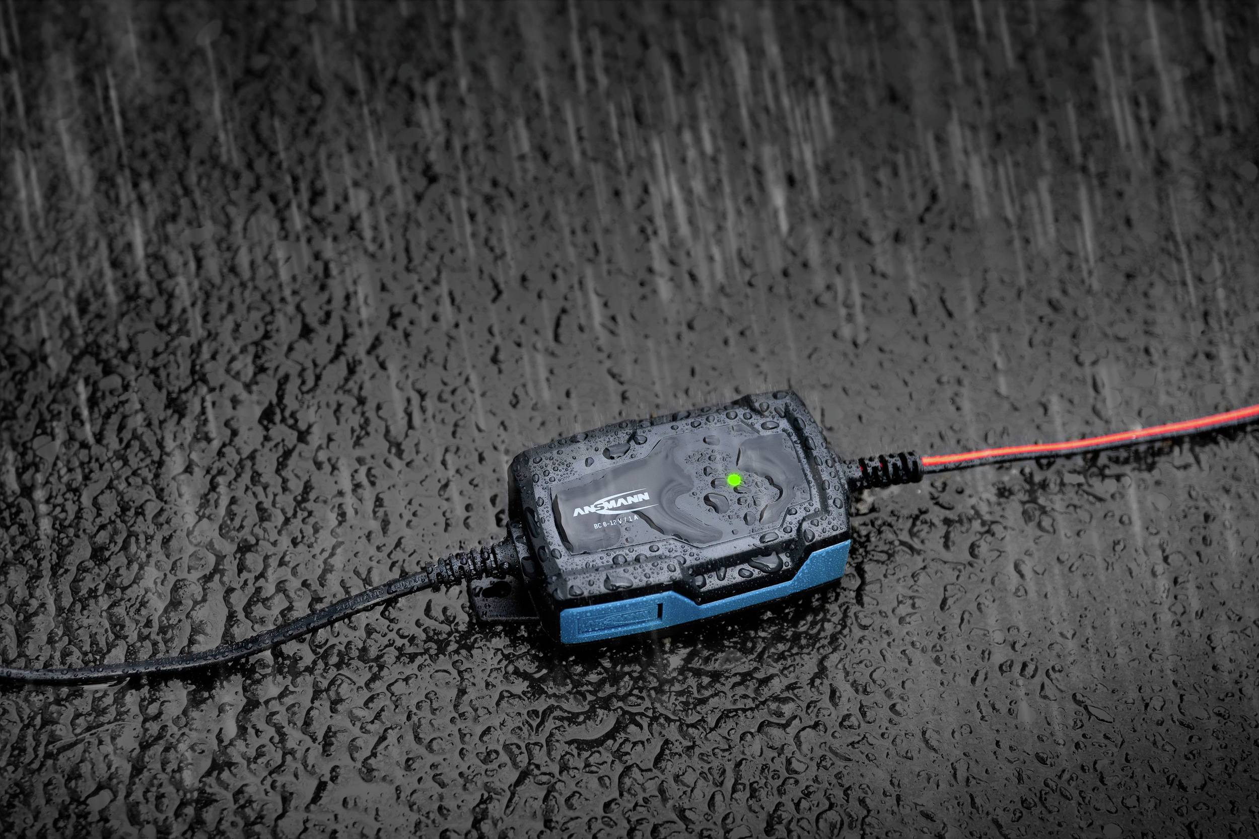 Een elektrisch apparaat op een natte ondergrond in de regen, toont een groene indicatielamp. Het geprofileerde patroon van de vloer is zichtbaar.