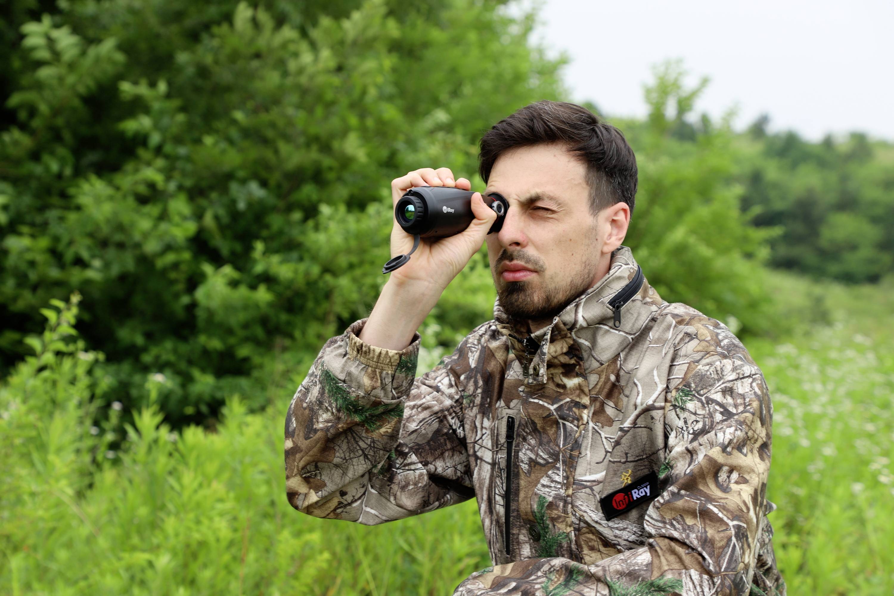 Een persoon in een camouflage-outfit gebruikt een monoculair om iets te observeren in een bosrijk gebied. Op de achtergrond staan dichte bomen.