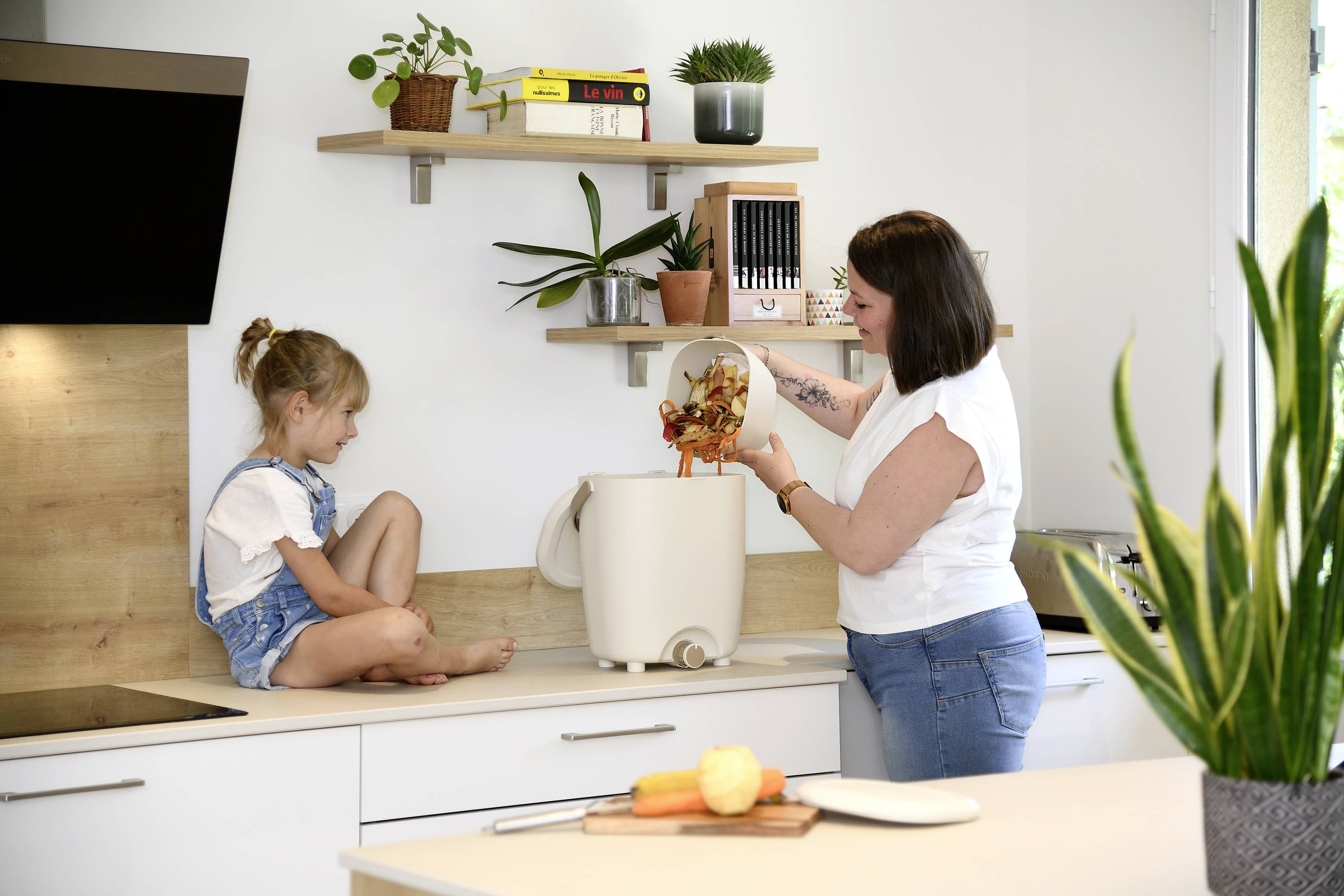 Vrouw gooit groenteafval in de keukencompostbak terwijl een klein meisje toekijkt. Moderne keukeninrichting met planten.