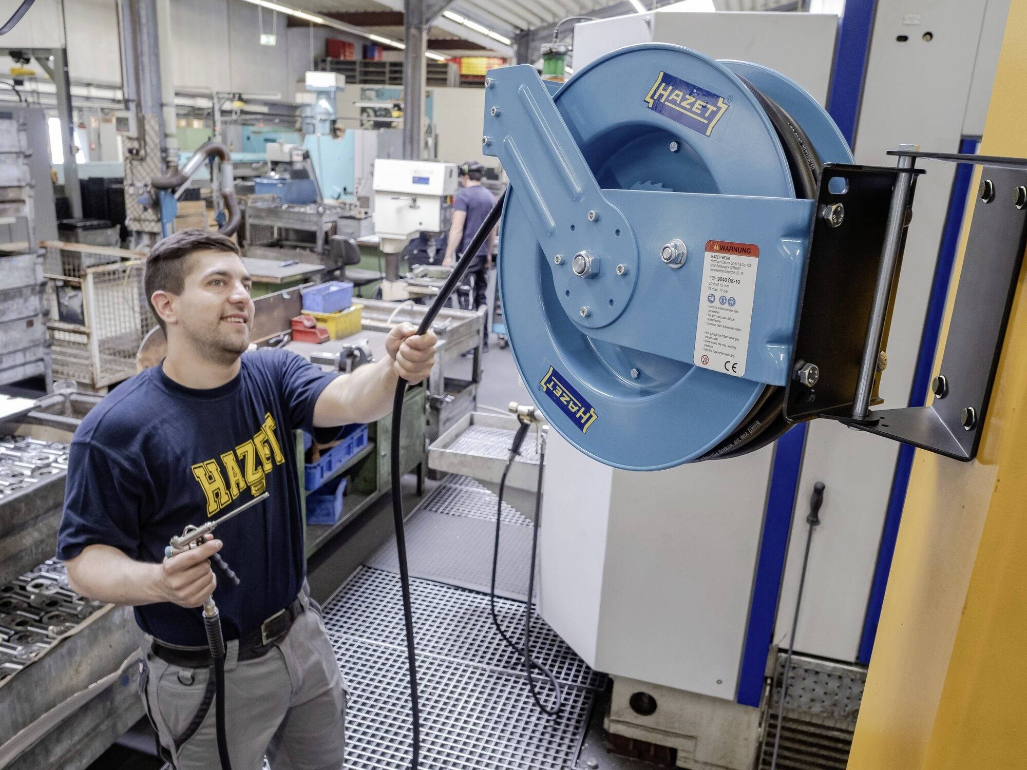 Een man in een werkplaats trekt een slang van een blauwe muurhaspel. Op de achtergrond zijn machines en gereedschappen te zien.