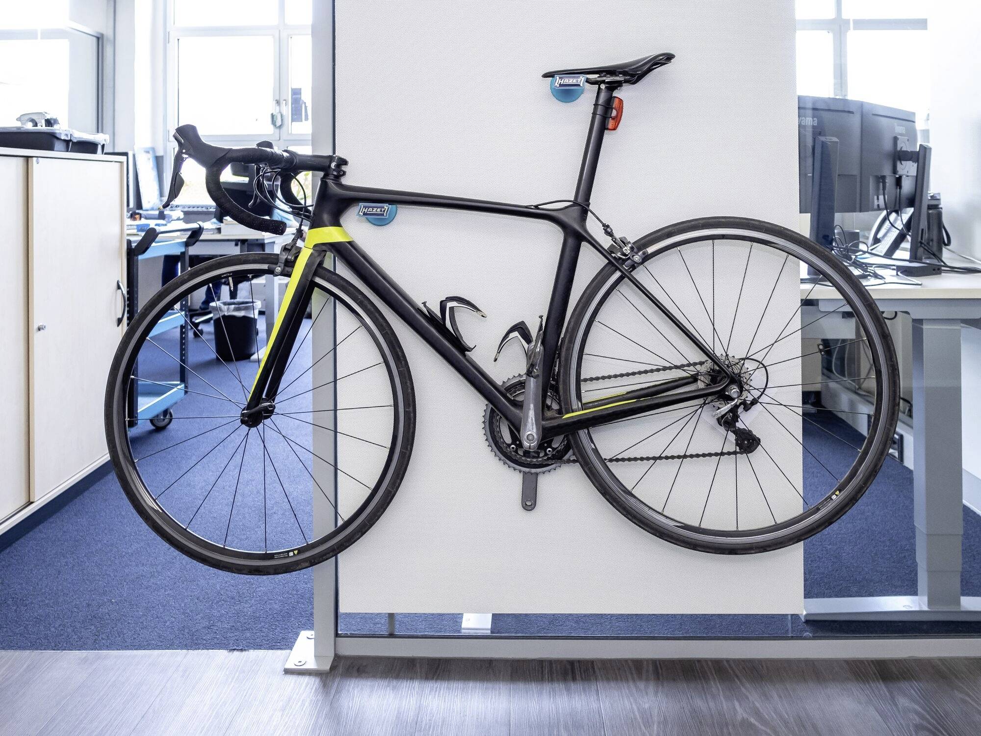 Een racefiets staat plaatsbesparend aan een wandsteun in een modern kantoor. Op de achtergrond zijn bureaus zichtbaar.