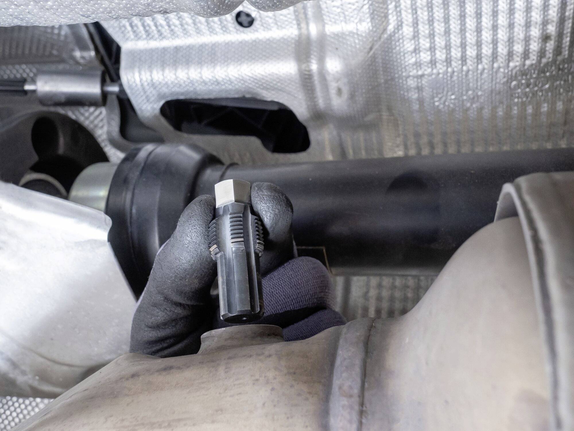 Persoon met een zwarte handschoen houdt een sensor onder een voertuig. De omgeving toont onderdelen van het uitlaatsysteem met metalen bekleding.