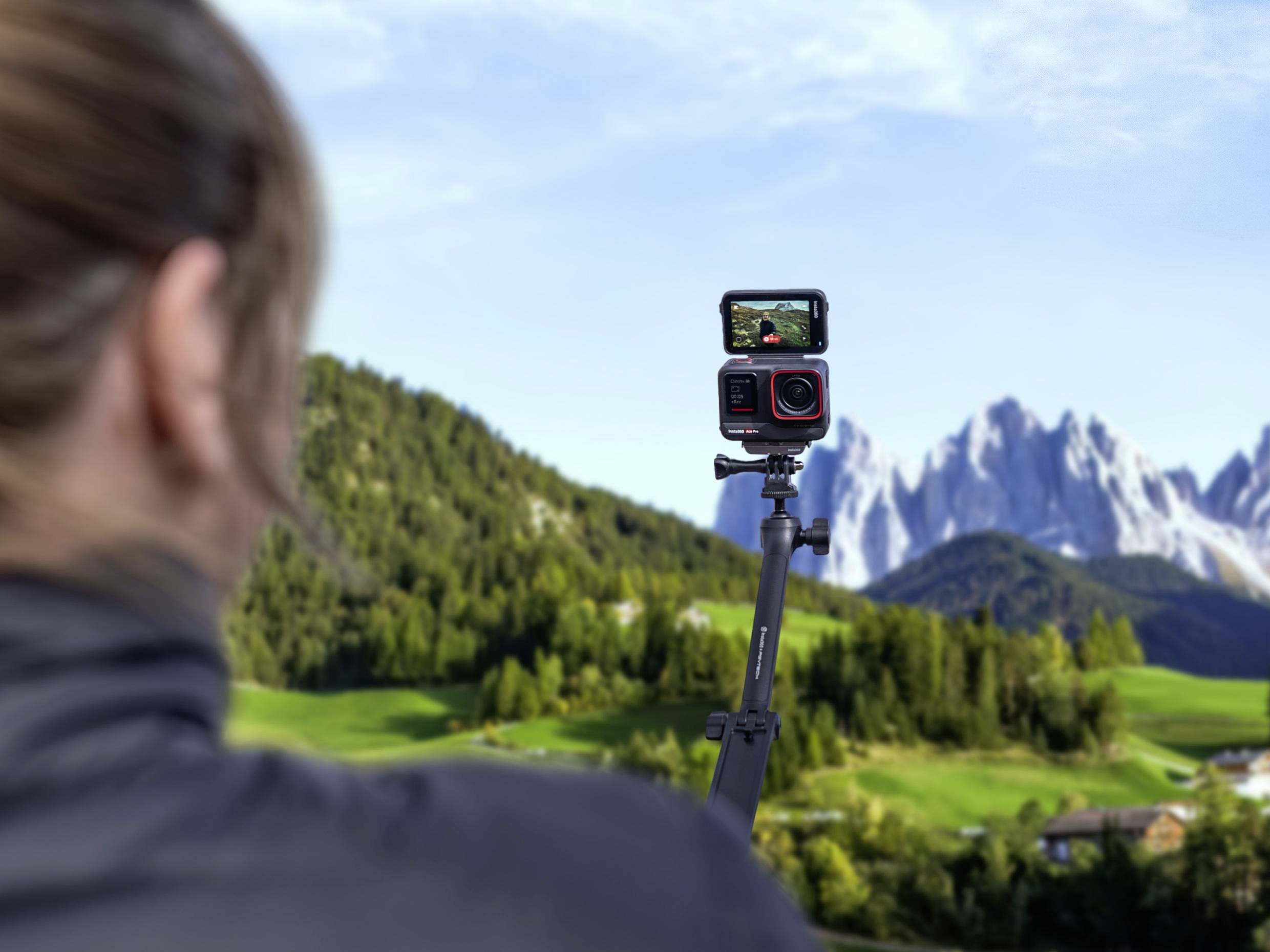 Een persoon filmt met een camera op een statief, gericht op een berglandschap. Op de achtergrond zijn bossen en bergen zichtbaar.