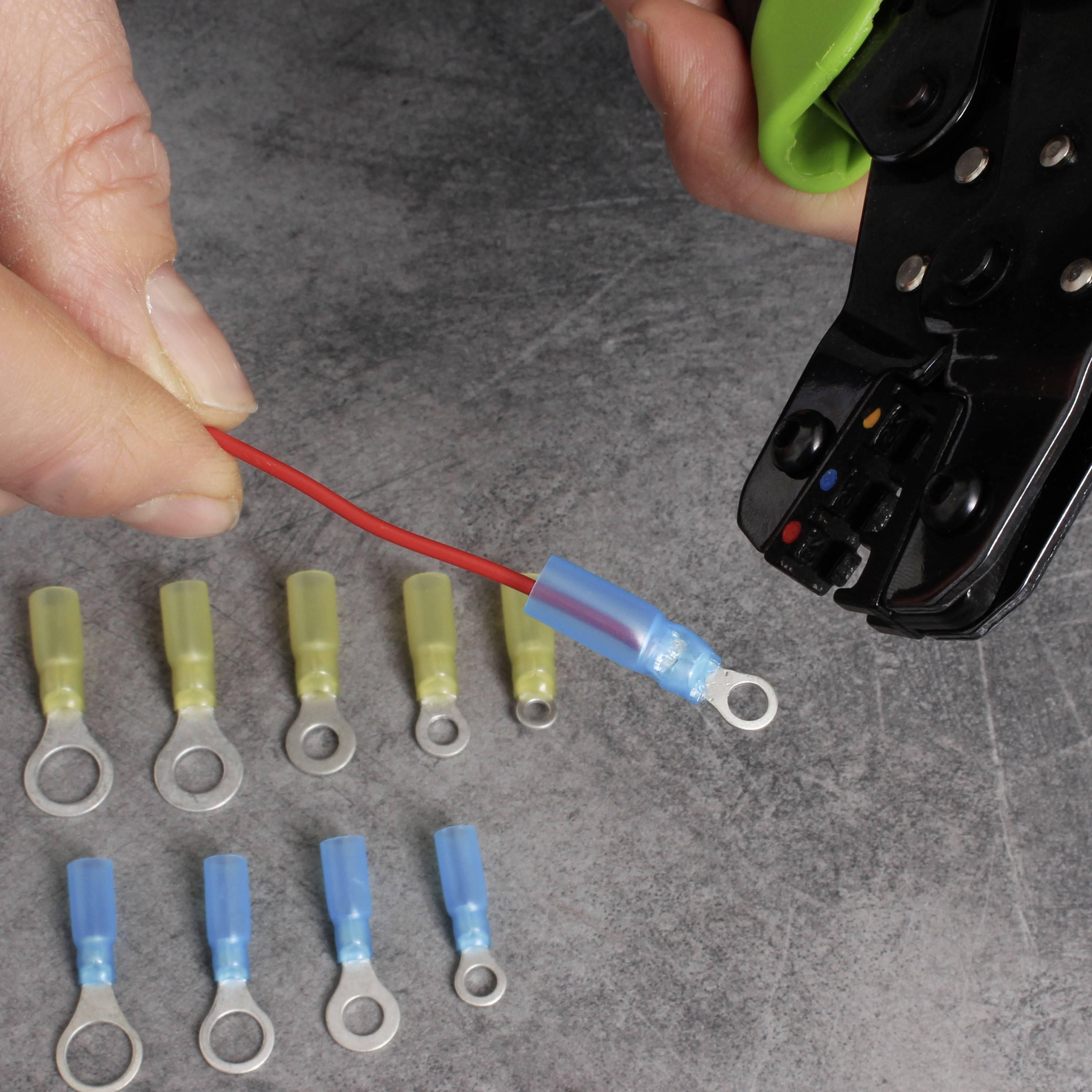 Een hand houdt een krimptang vast die een rode draad afknipt; ernaast liggen verschillende kabelschoenen op een grijze ondergrond.