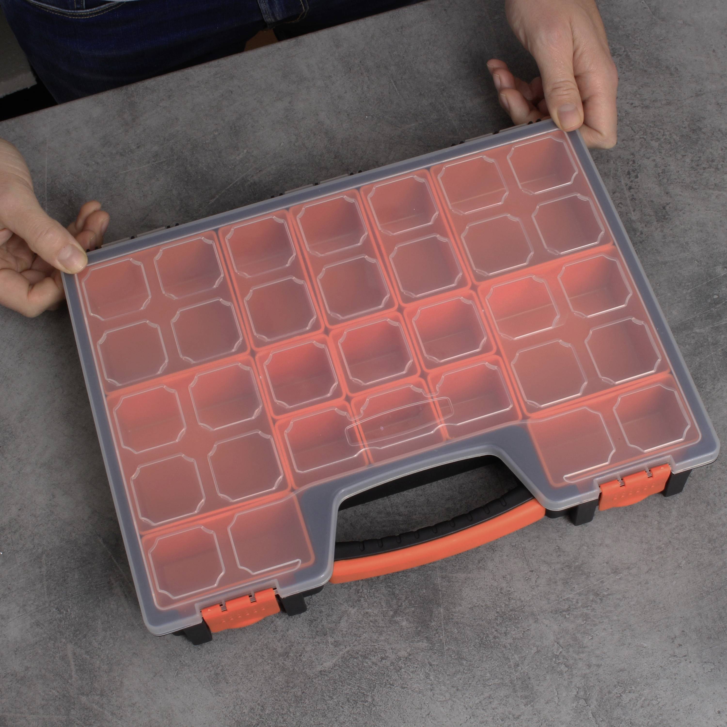 Een hand houdt een transparante gereedschapskist met een oranje handgreep en meerdere compartimenten. De kist staat op een grijze ondergrond.