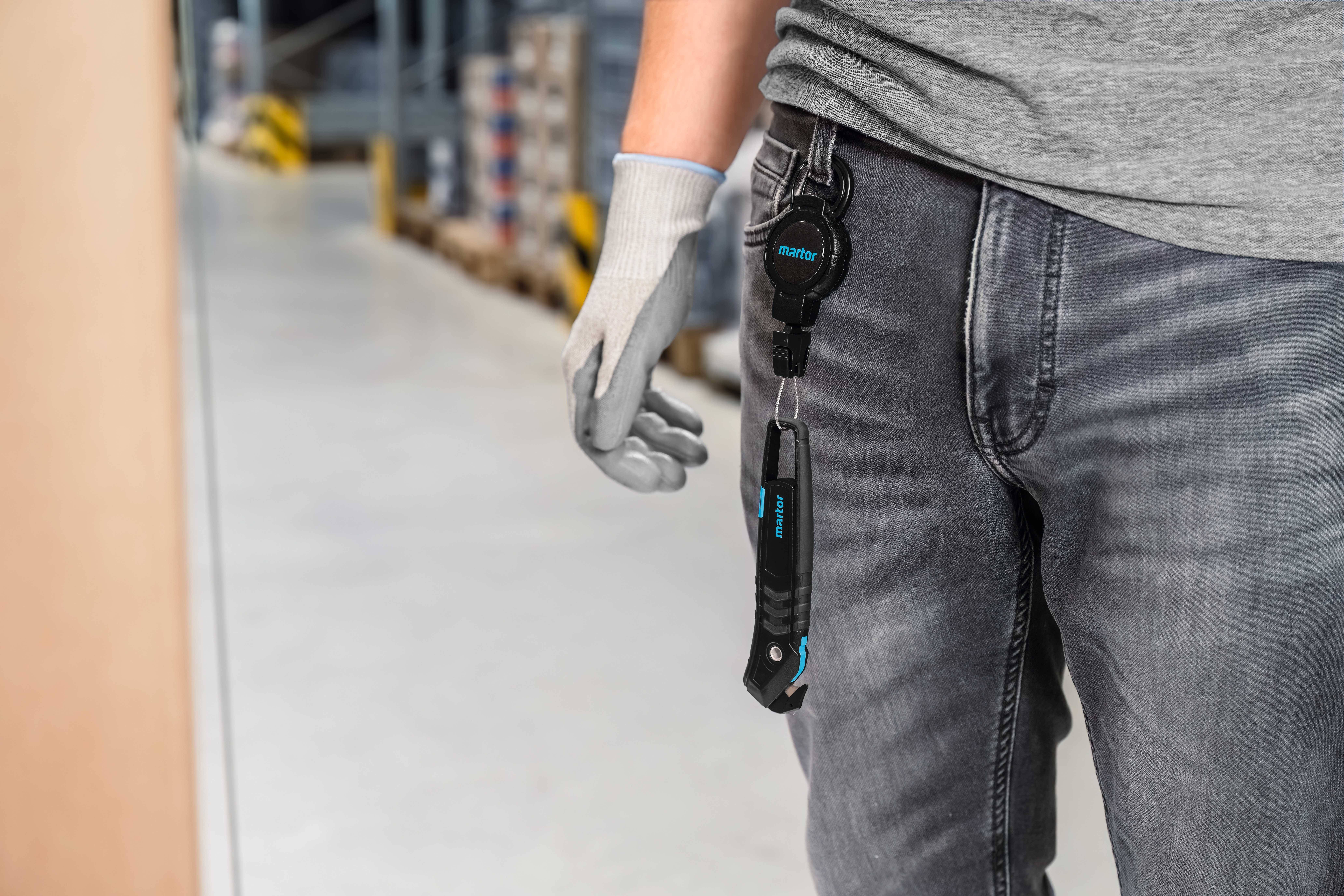 Een persoon in het magazijn draagt een riem met daaraan bevestigde gereedschappen. Ze draagt grijze handschoenen en een spijkerbroek. De achtergrond toont stellingen en dozen.