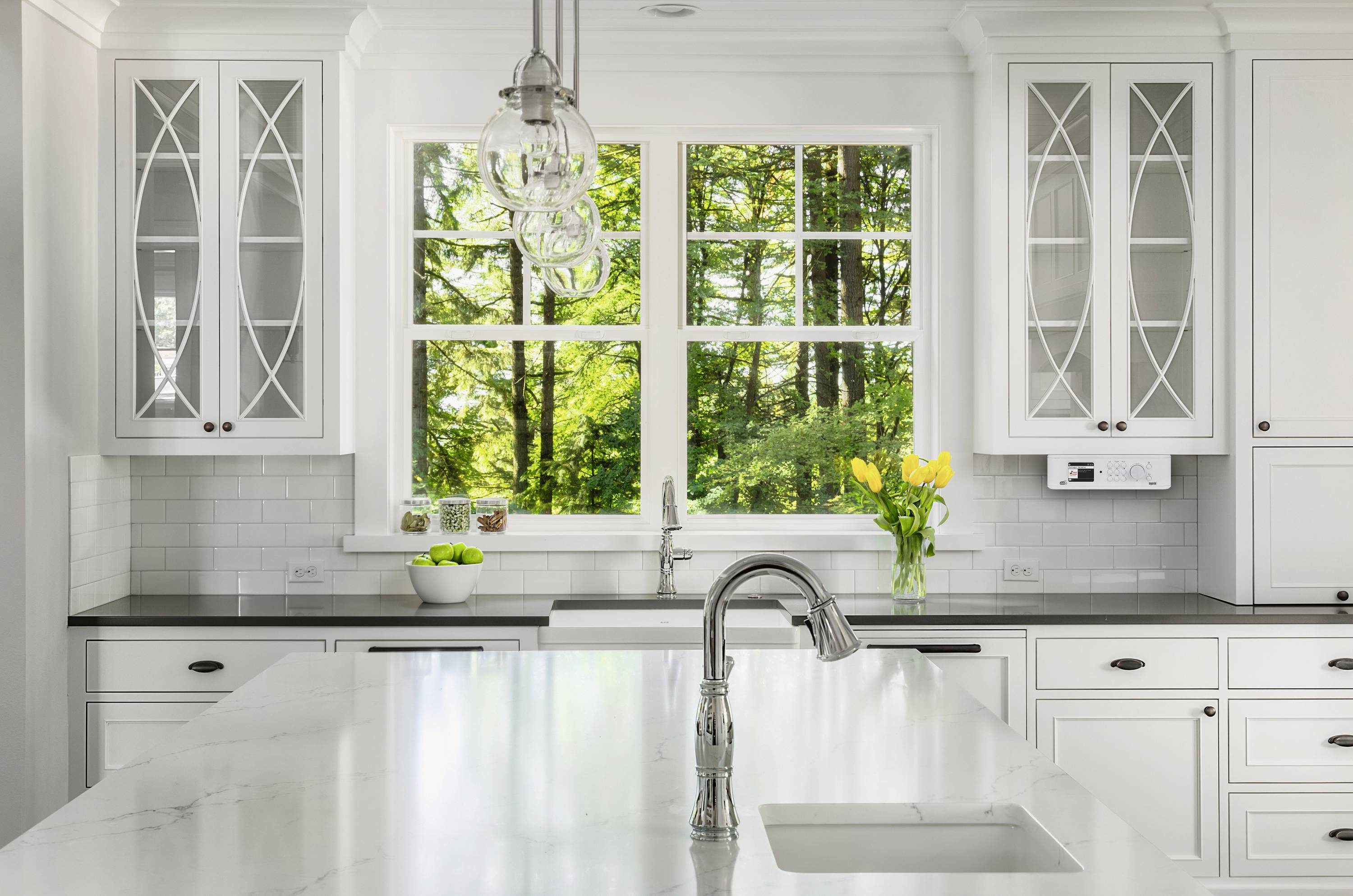 Elegante, moderne keuken met witte uitrusting en grote raampartij. Buiten is een groene bos zichtbaar. Vaas met gele tulpen op het aanrecht.