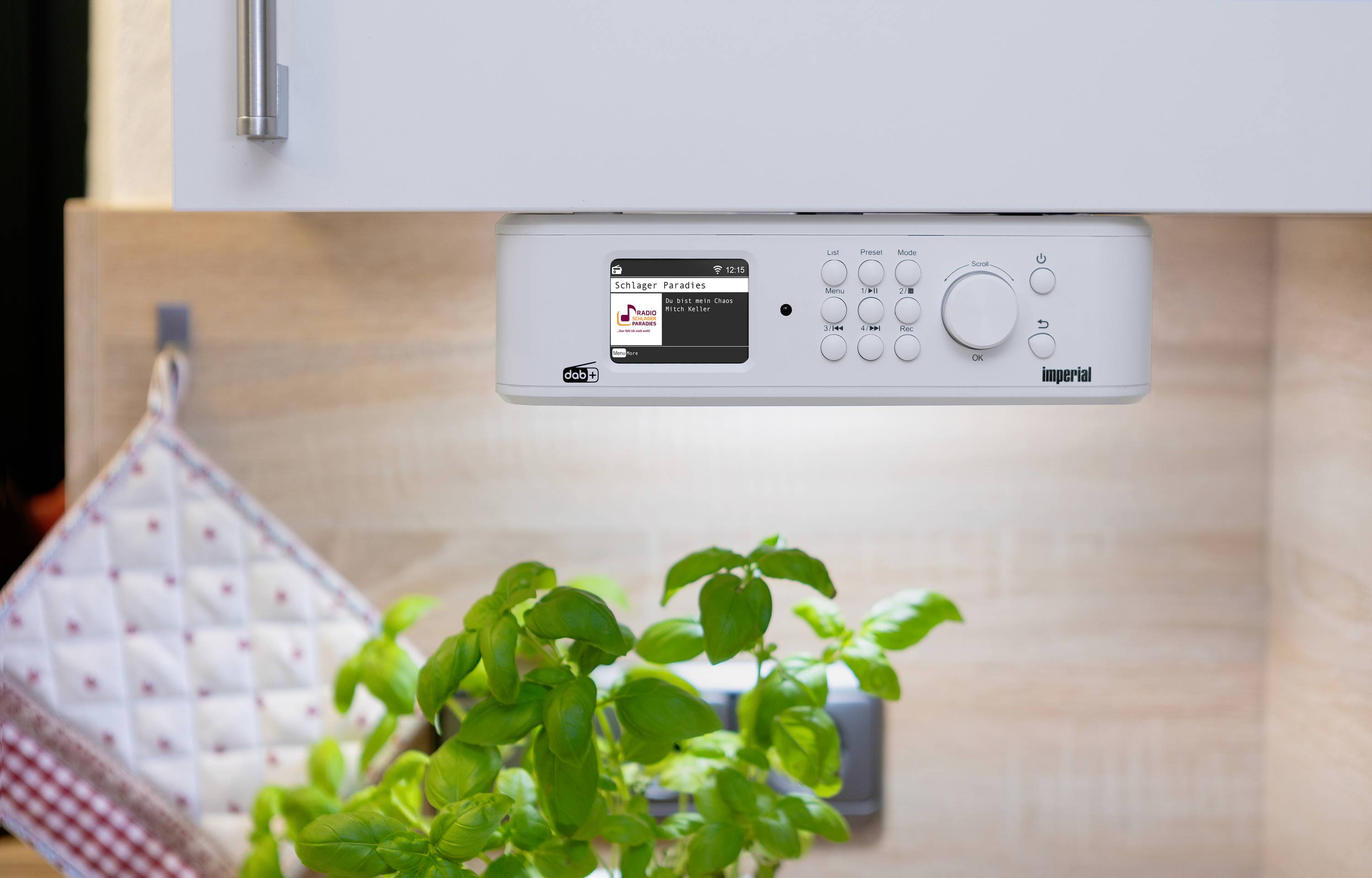 Onder een keukenkast is een wit keukenradio met display bevestigd. Eronder staat een groene plant op het aanrecht.