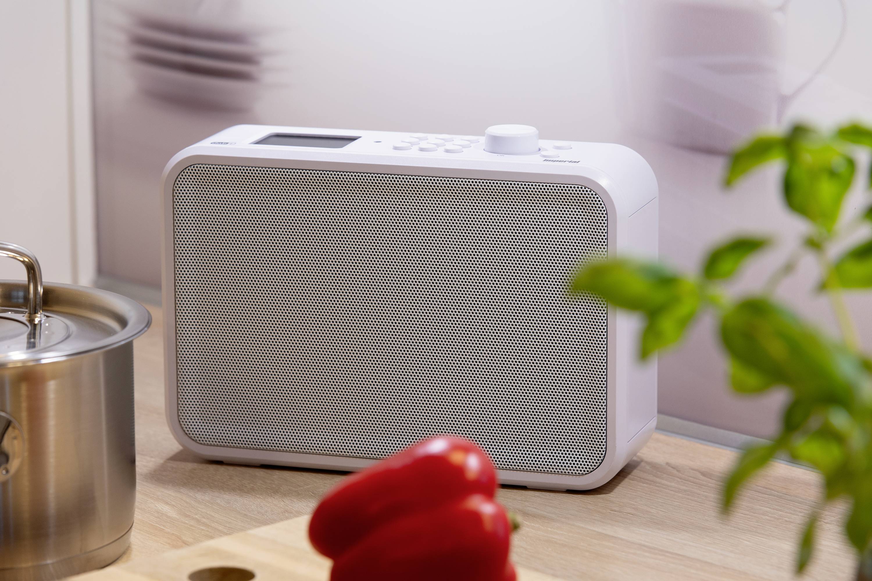 Een wit radio staat op een aanrecht naast een rode paprika en een potplant, op de achtergrond wazig een pan.