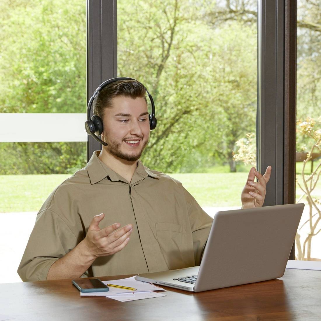 Een man zit glimlachend voor een laptop, draagt koptelefoon, gebaren makend met zijn hand, omringd door een groene tuin door het raam.