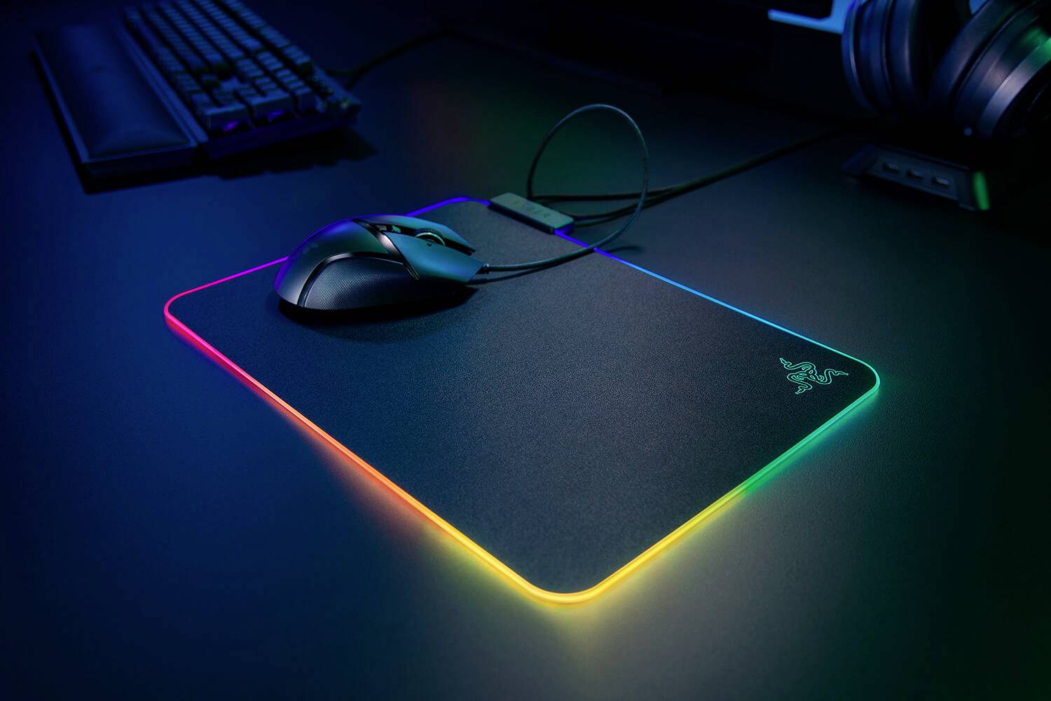 Een gamingmuis ligt op een verlicht muismat met RGB-led's, geplaatst in een donkere ruimte naast een toetsenbord en koptelefoon.