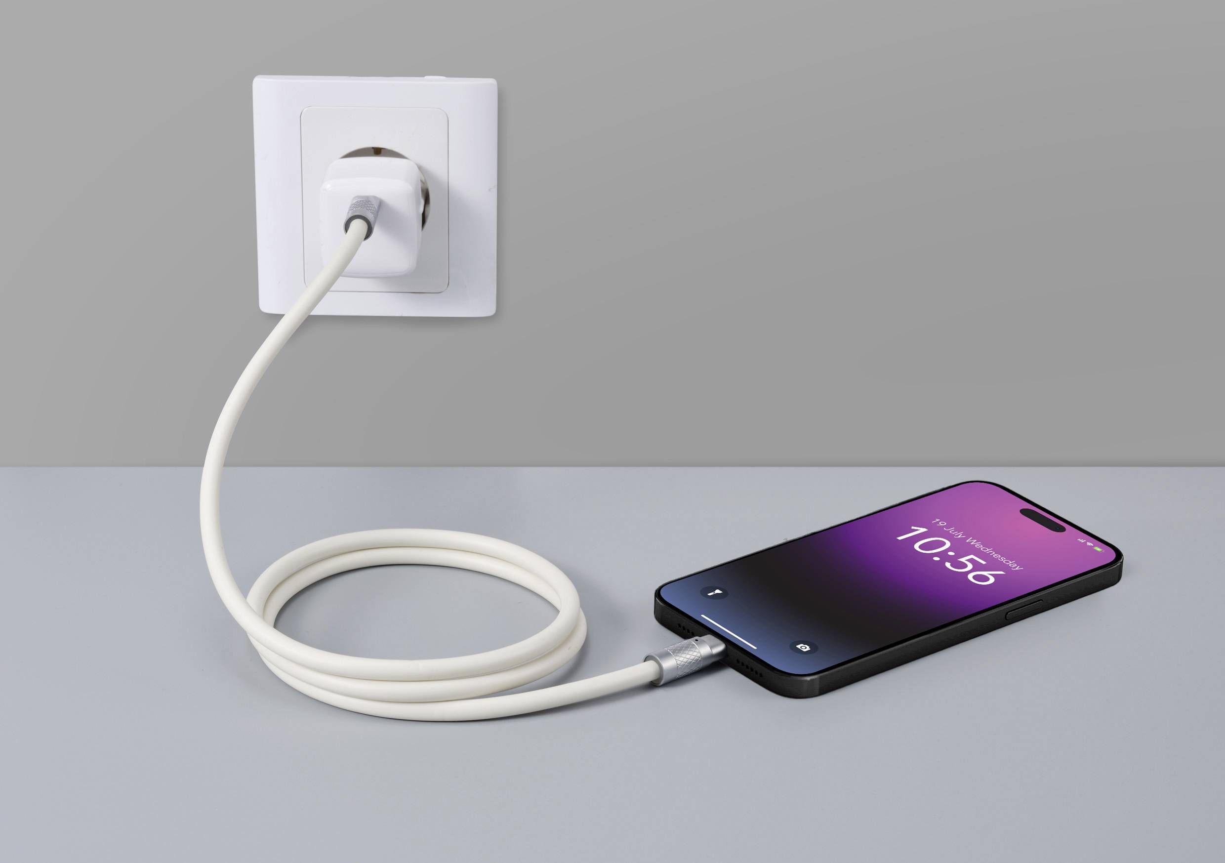 Een smartphone wordt opgeladen met een spiraalvormig oplaadkabel bij een stopcontact aan de muur. Het scherm toont de tijd 10:56.