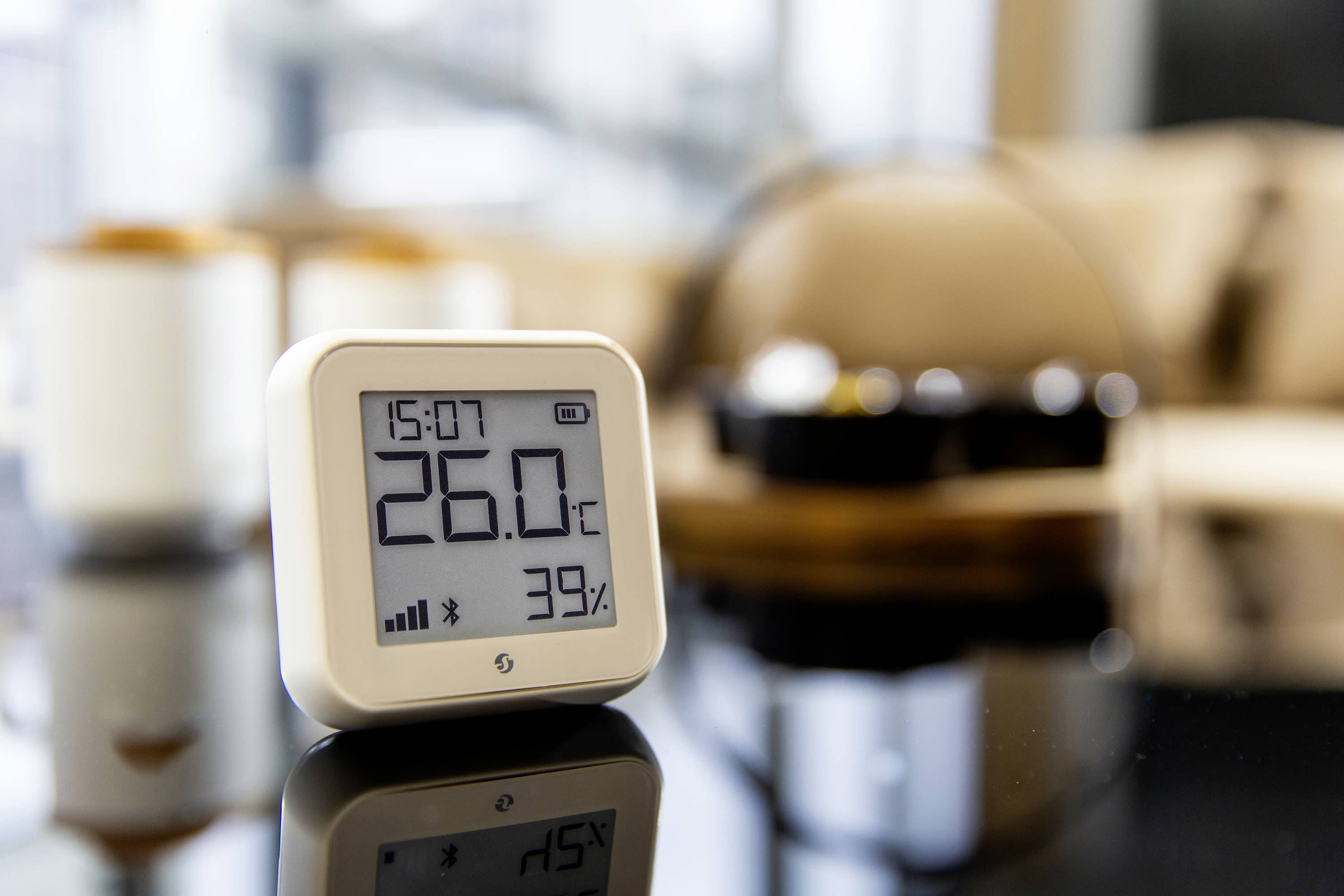 Een digitale thermometer op een tafel toont 26,0°C en 39% luchtvochtigheid. Op de achtergrond zijn wazig woonkamermeubilair te zien.