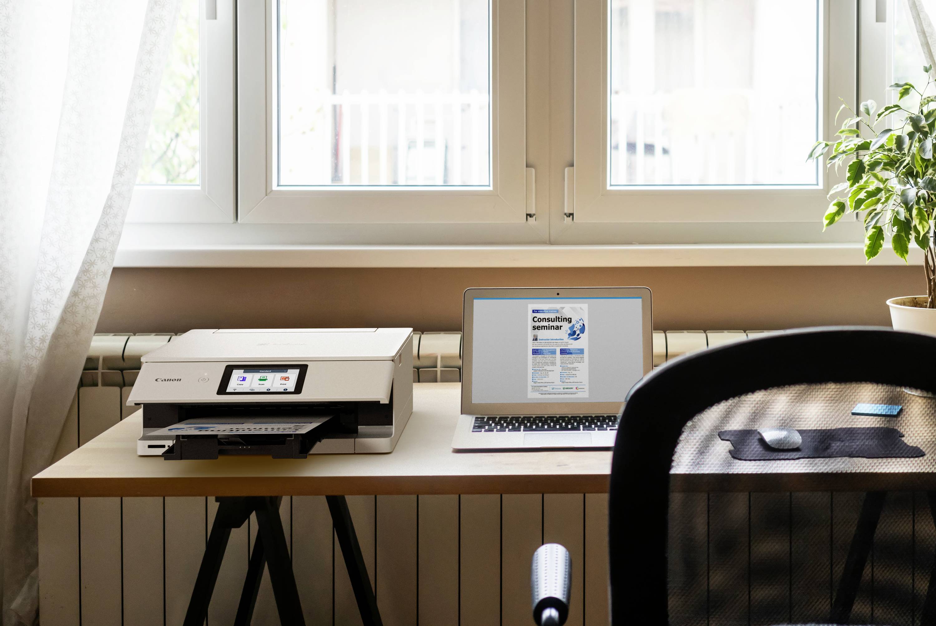 Een bureau met een geopende laptop waarop een document zichtbaar is, naast een printer en een kamerplant. Op de achtergrond een raam.