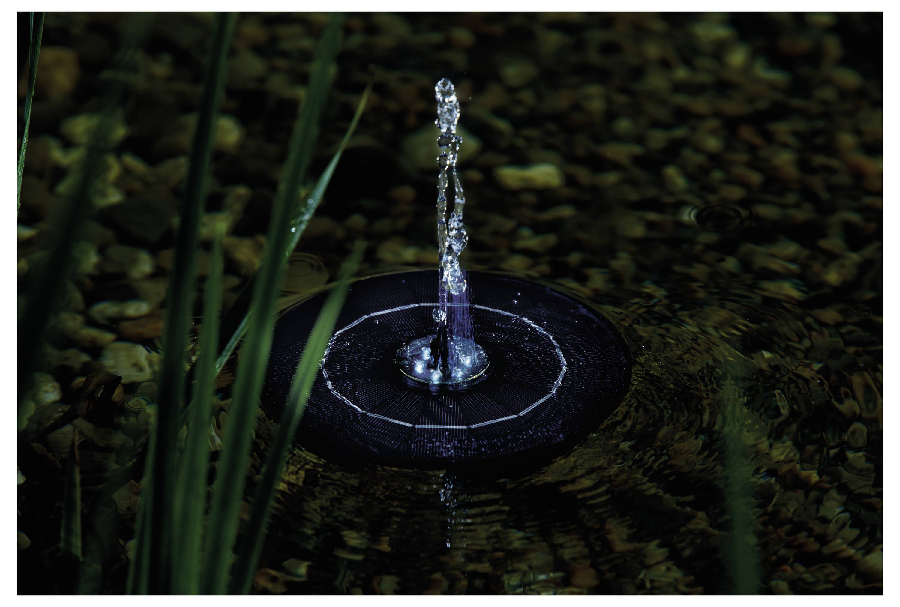 Een kleine zonneënergie-fontein met een waterstraal die in het midden omhoog rijst, omringd door kiezels en groen gras dat weerkaatst in het water.