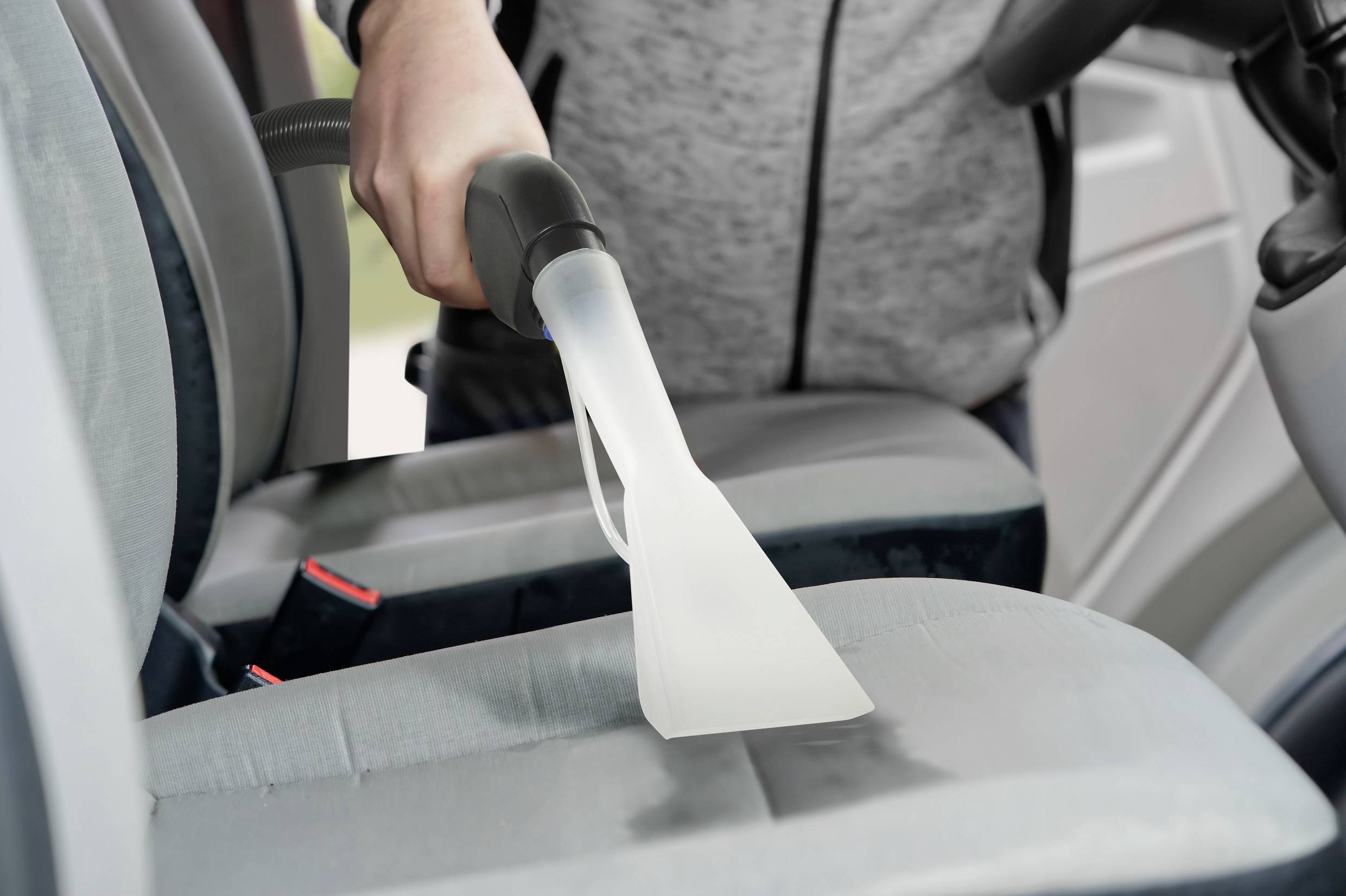 Een persoon reinigt de autostoel met een nat-zuiger. De grijze stoel wordt ontdaan van vlekken. De scène geeft een gevoel van reinheid.
