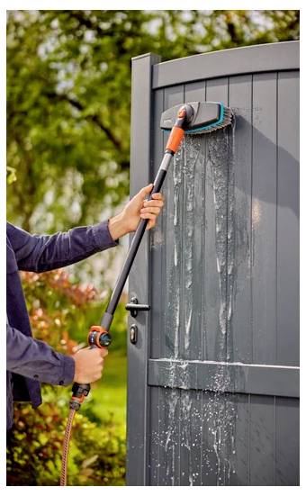 Een persoon gebruikt een langgesteelde borstel en slang om een grijze buitenpoort schoon te maken, waarbij water over het oppervlak stroomt in een tuinomgeving.