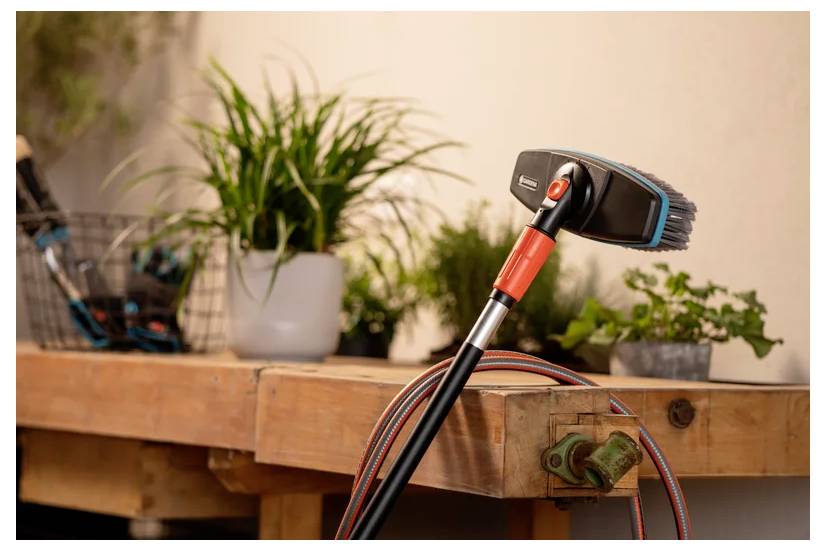 Een schoonmaakborstel met een rode steel ligt op een houten werkbank omringd door potplanten, waardoor een nette, tuinieren-geïnspireerde sfeer wordt gecreëerd.