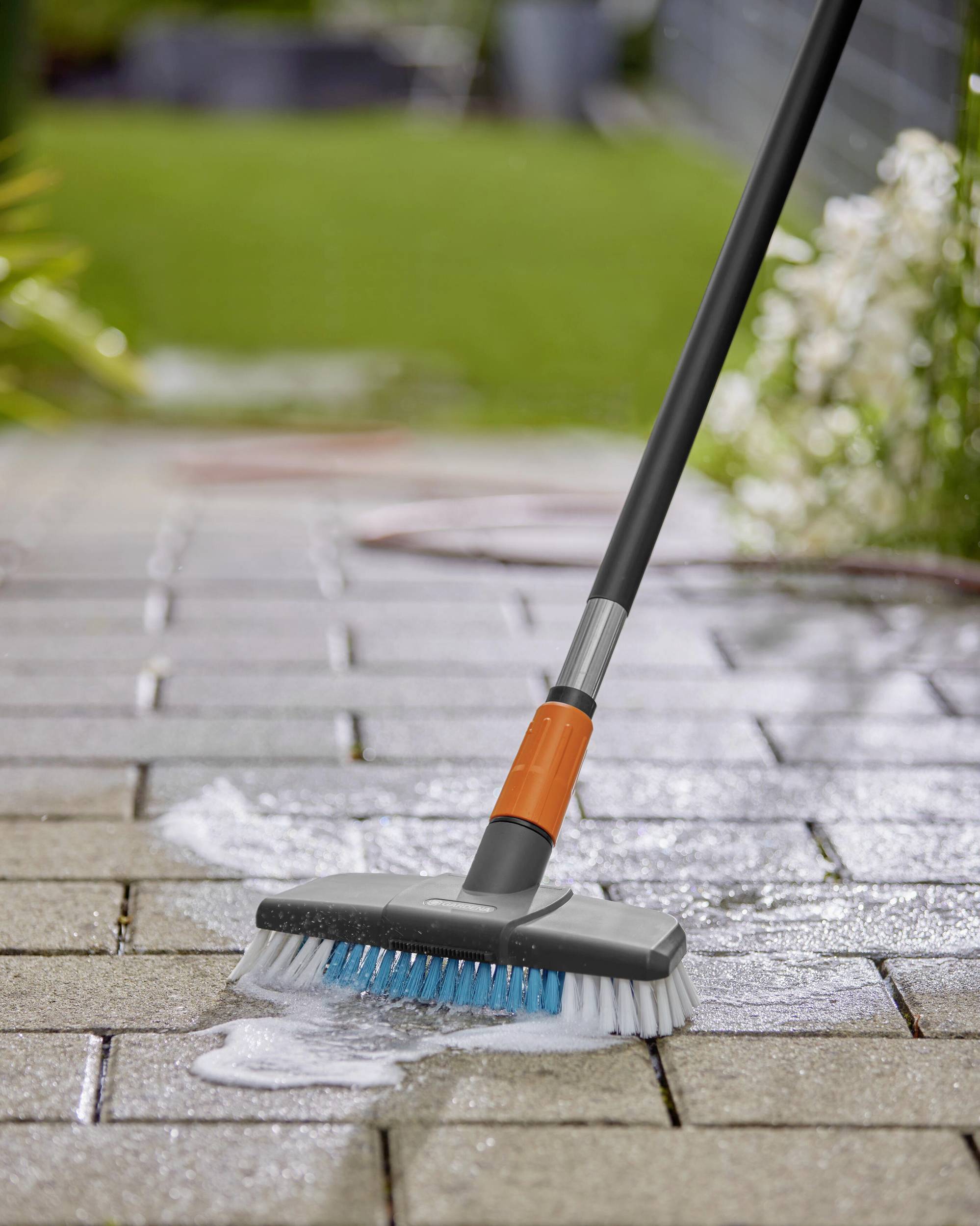 Een schoonmaakbezem met een oranje steel scrubt een bestrate terras. Zeepwater vormt schuimende sporen op de tegels.