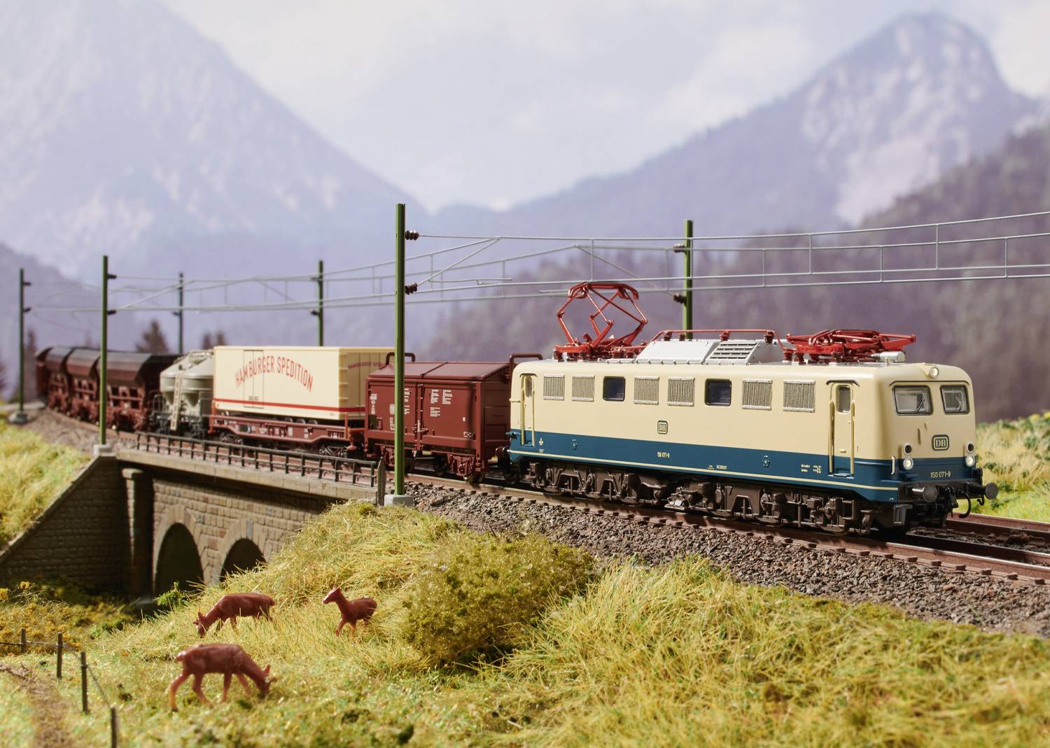 Een modeltrein met goederenwagons rijdt over een brug in een landschap met bergen op de achtergrond en wild dat graast op de voorgrond.