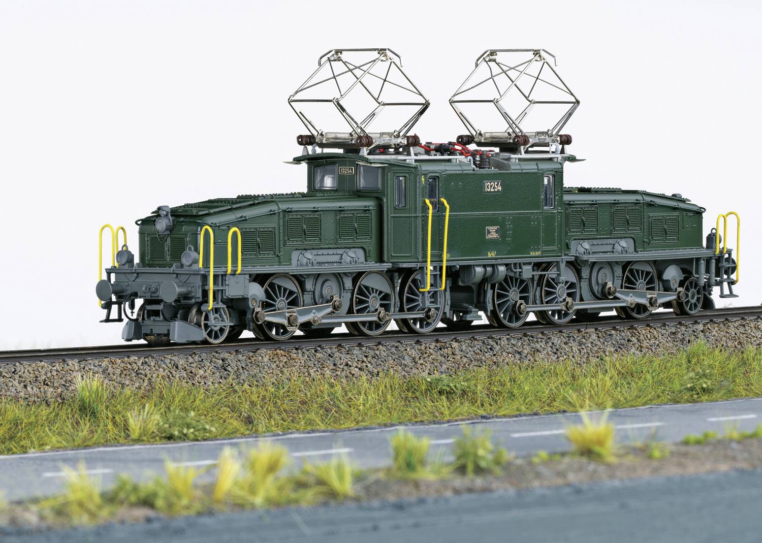 Een groen, gedetailleerd model van een oude elektrische locomotief met meerdere pantografen staat op een spoor, omgeven door gras.