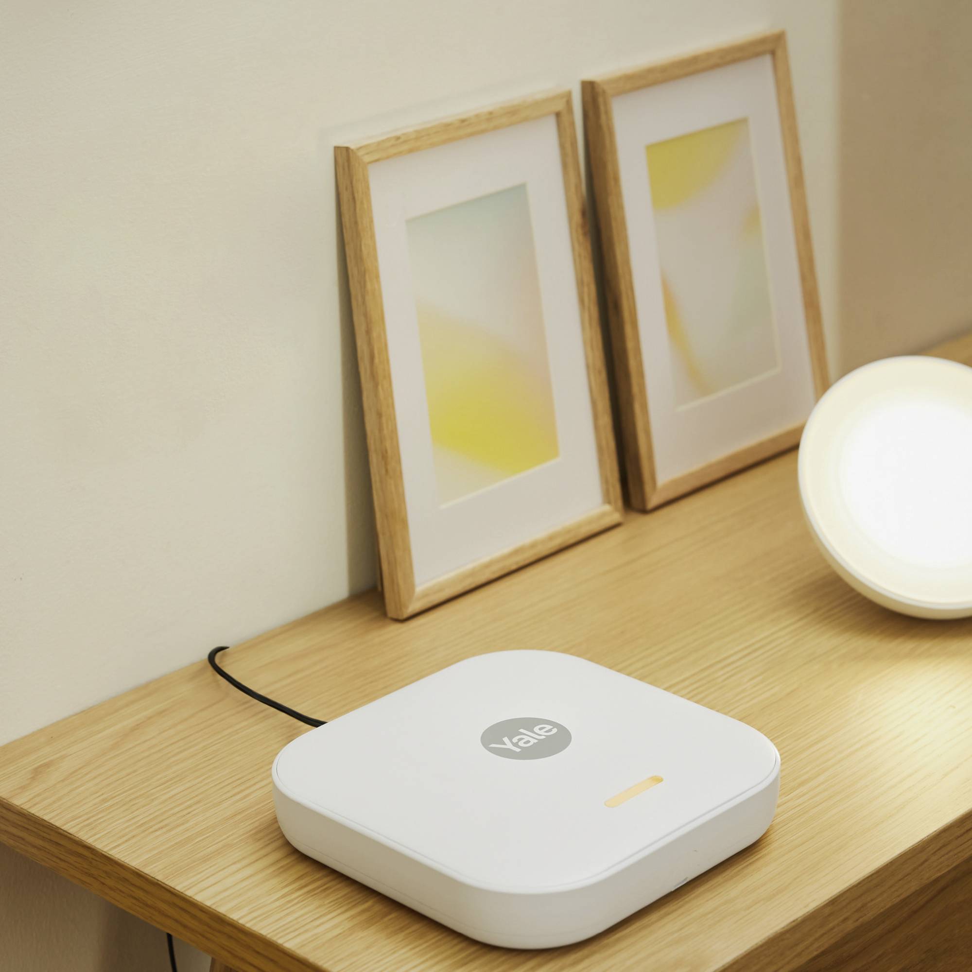 Een wit Smart Home-apparaat met 'Yale'-logo op een houten tafel, naast twee ingelijste afbeeldingen en een lamp.