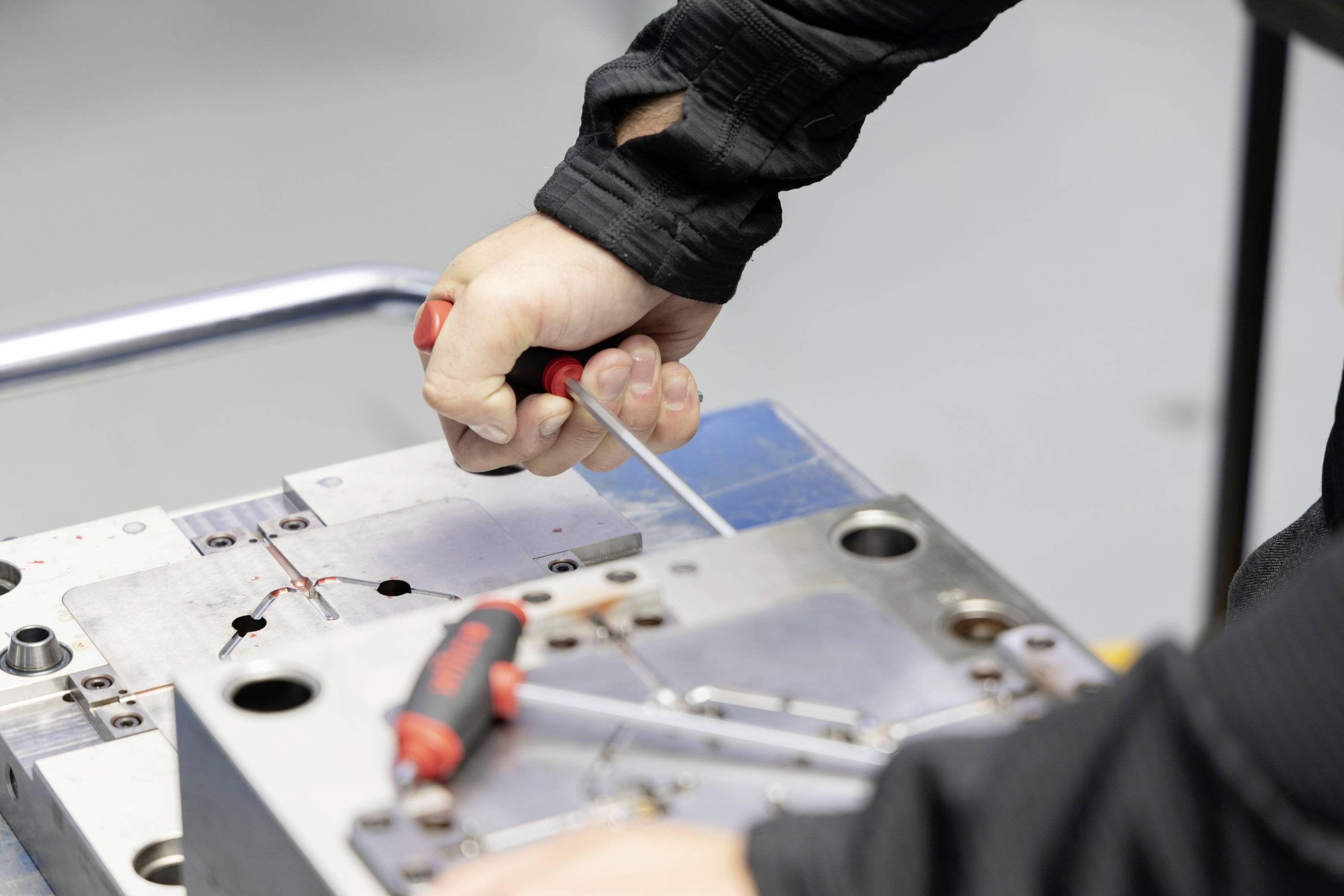 Een persoon houdt een schroevendraaier vast en werkt aan een metalen gereedschap op een tafel. Gereedschappen en schroevendraaiers liggen ernaast.