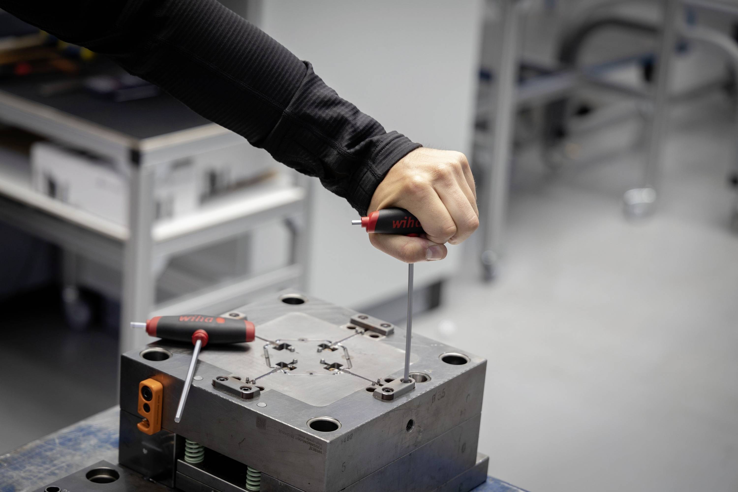 Een persoon monteert een metalen onderdeel met twee schroevendraaiers in een werkplaatsomgeving.