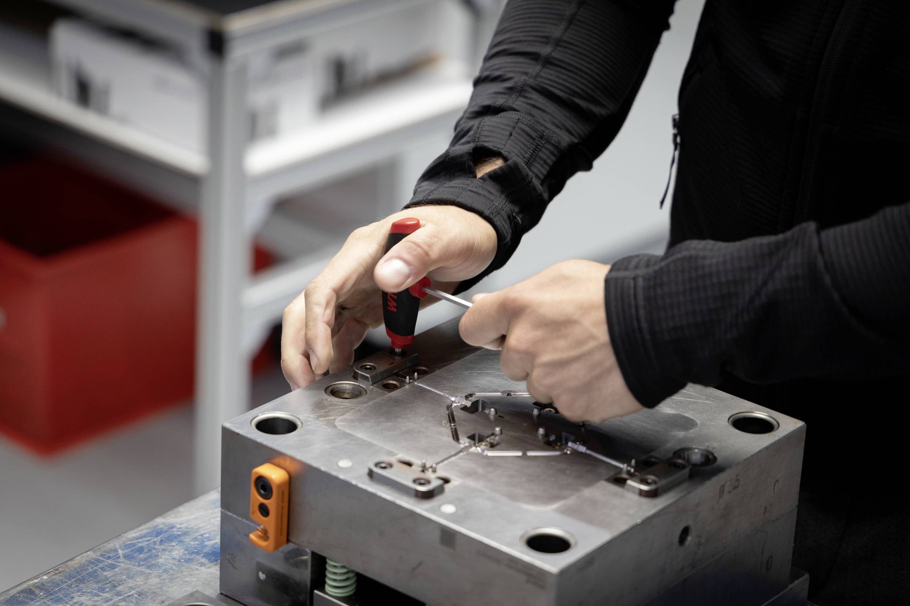Een persoon werkt aan een metalen gereedschap of vorm met een schroevendraaier in een werkplaatsomgeving.
