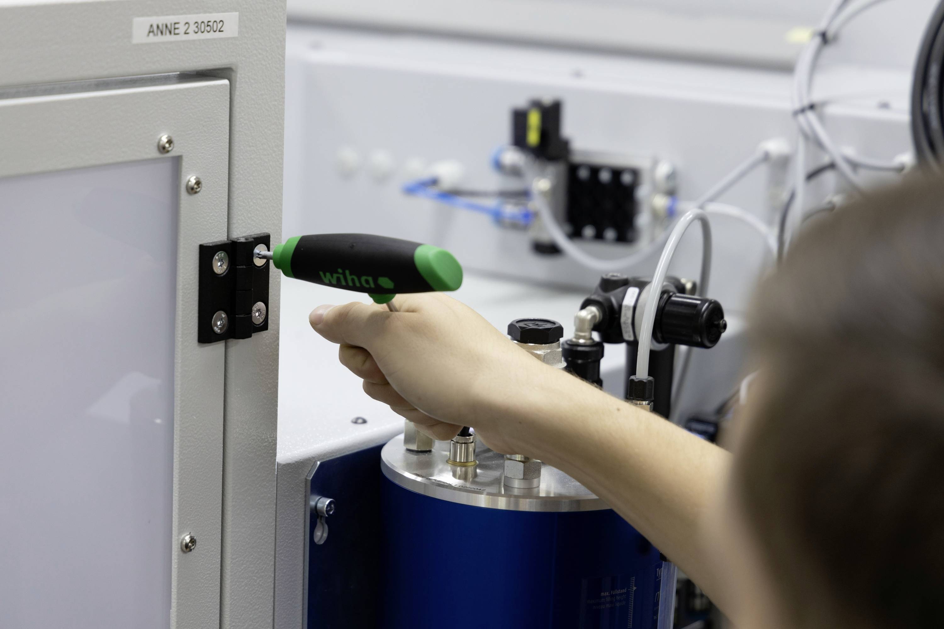 Een persoon draait een schroef vast met een groene schroevendraaier aan een machinedeur. Op de achtergrond zijn technische apparaten zichtbaar.