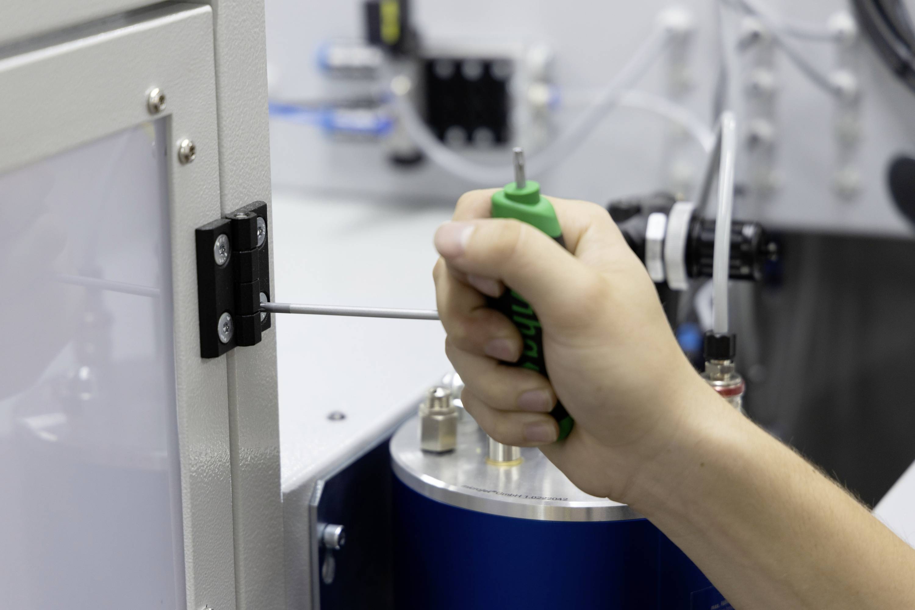 Een hand houdt een schroevendraaier vast en draait een schroef vast aan een machinebehuizing. Op de achtergrond zijn machinedelen zichtbaar.