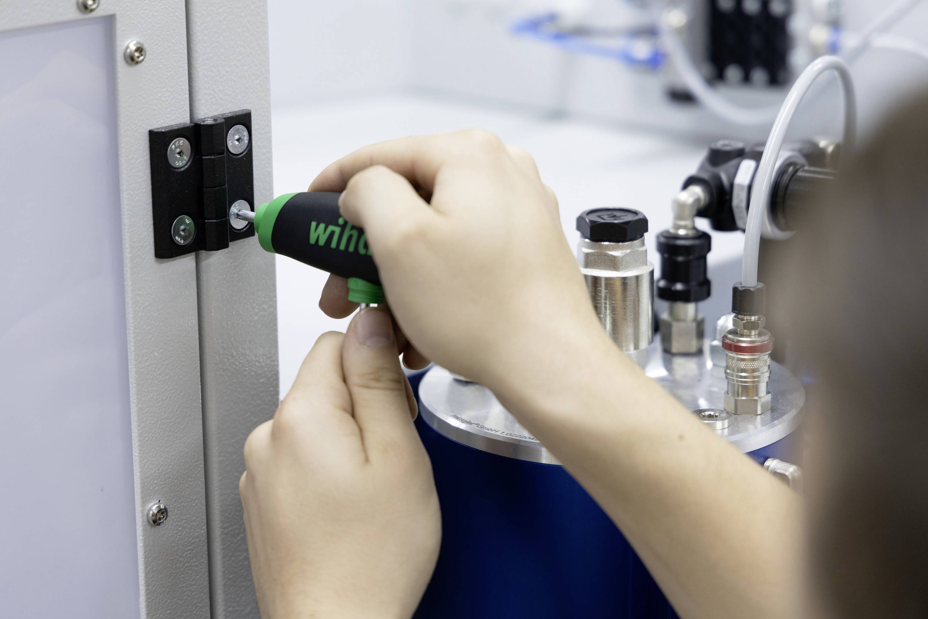 Een persoon draait een schroef vast in een metalen behuizing met een schroevendraaier. Op de achtergrond zijn verschillende machinedelen zichtbaar.