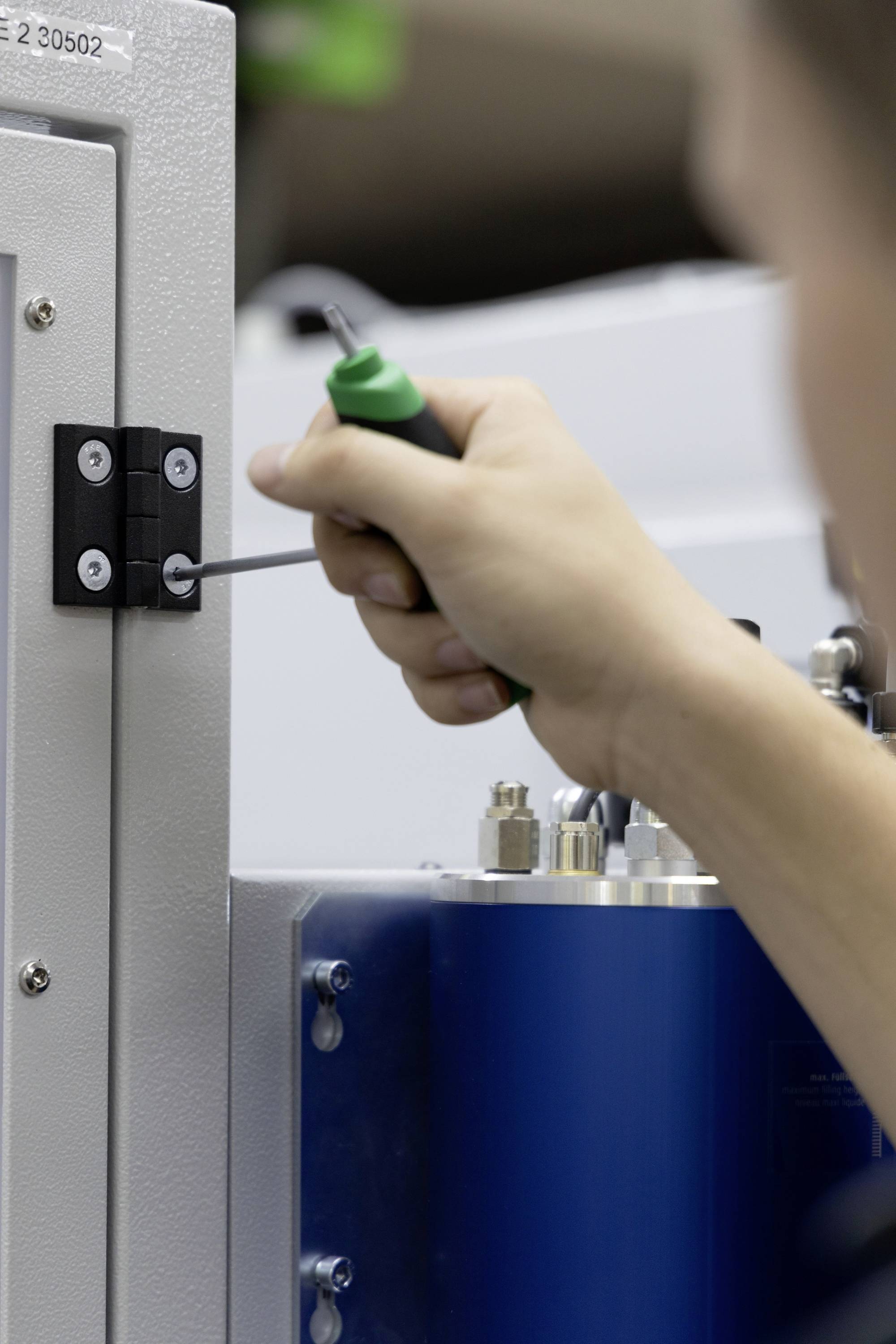 Persoon repareert apparaat met schroevendraaier. Verricht technische werkzaamheden aan een metalen kist met scharnier. Geconcentreerd bezig met gedetailleerd werk.