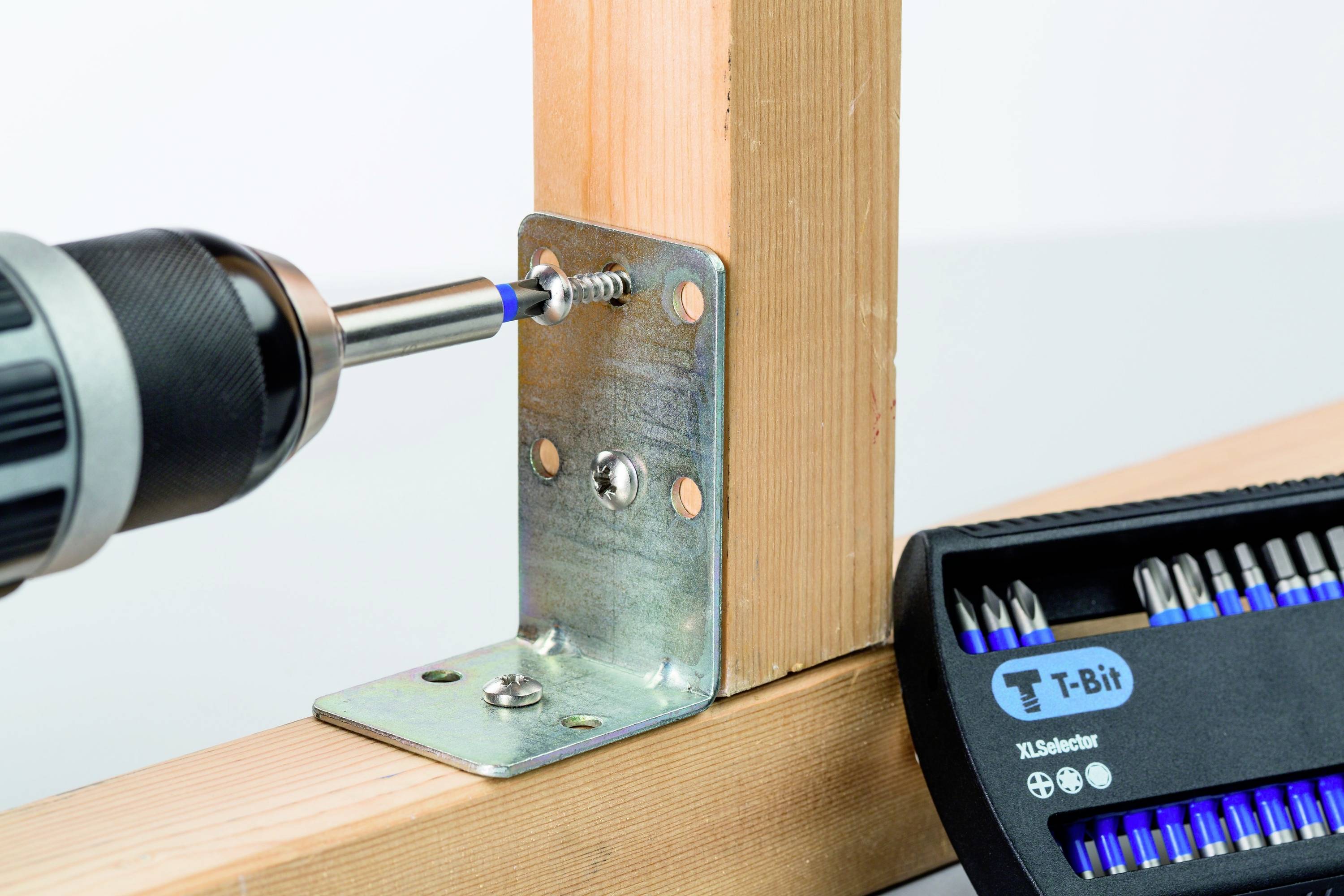 Een boormachine bevestigt een schroef in een metalen beugel die twee houten platen met elkaar verbindt. Ernaast ligt een set schroevendraaierbitjes.