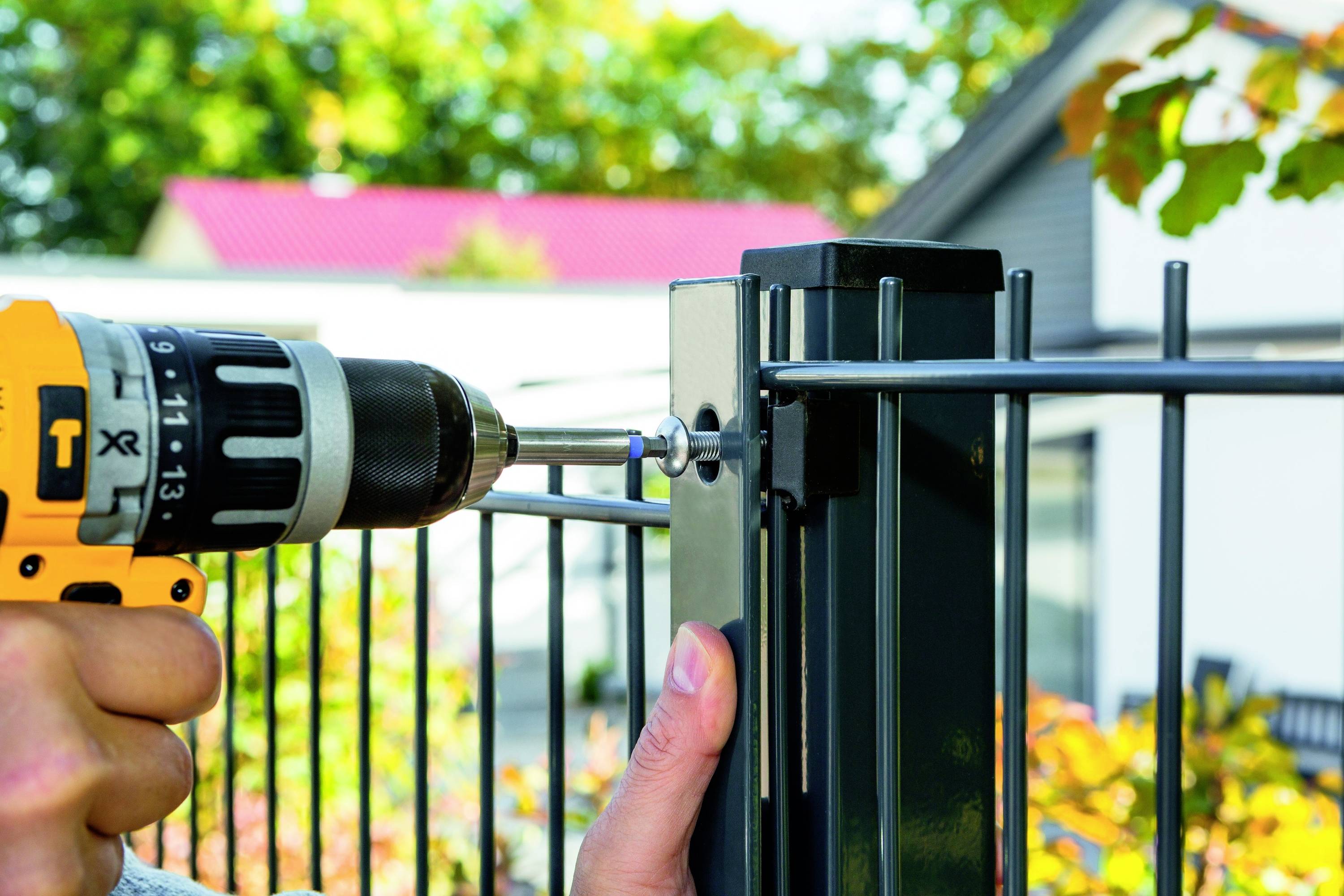 Een hand houdt een accuschroefmachine vast die een schroef in een metalen hekpaal draait. Op de achtergrond zijn bomen en een huis zichtbaar.
