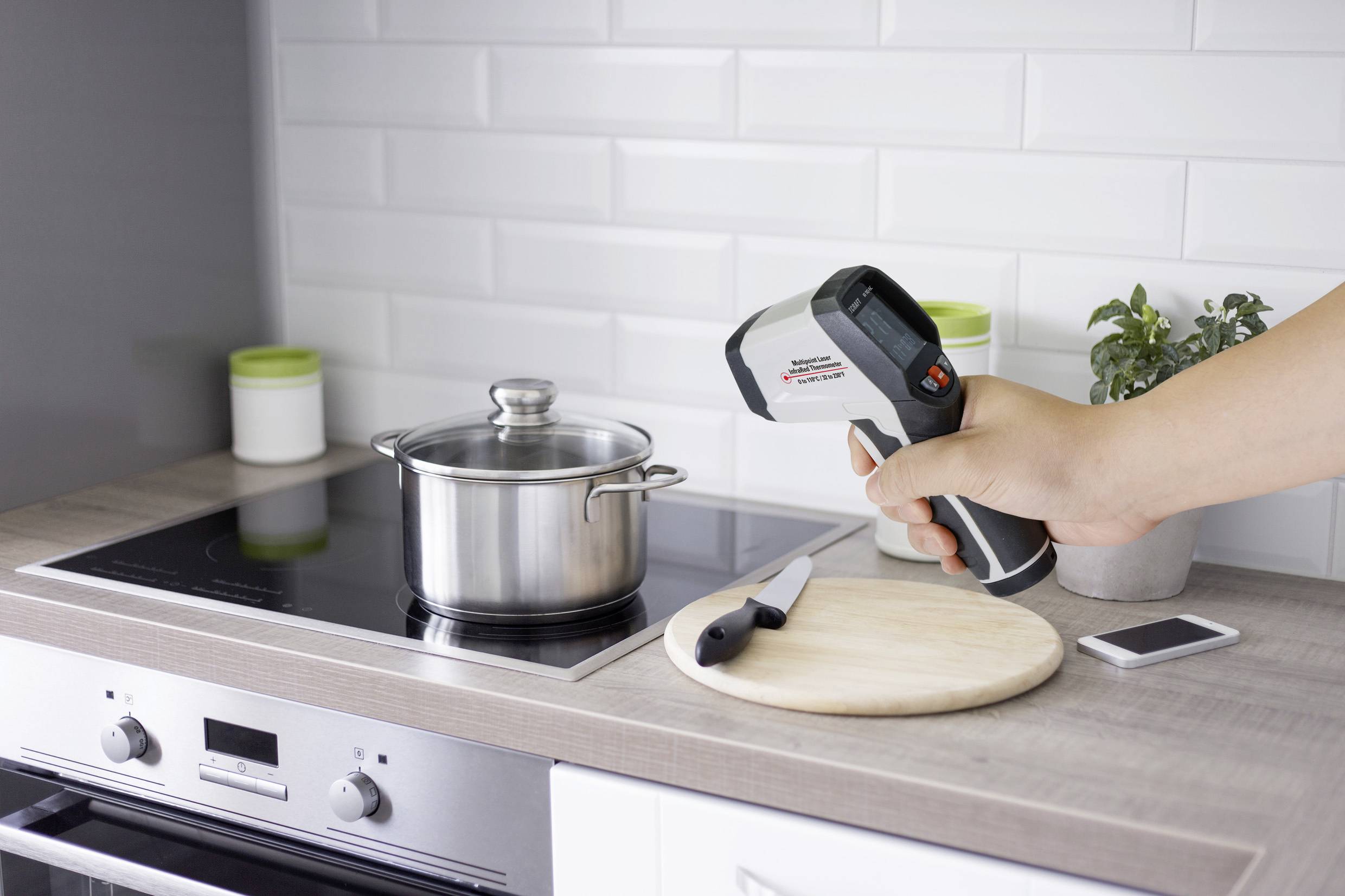 Een hand houdt een infrarood-thermometer vast die wijst naar een pan op een elektrisch fornuis. Ernaast liggen een mes en een snijplank.