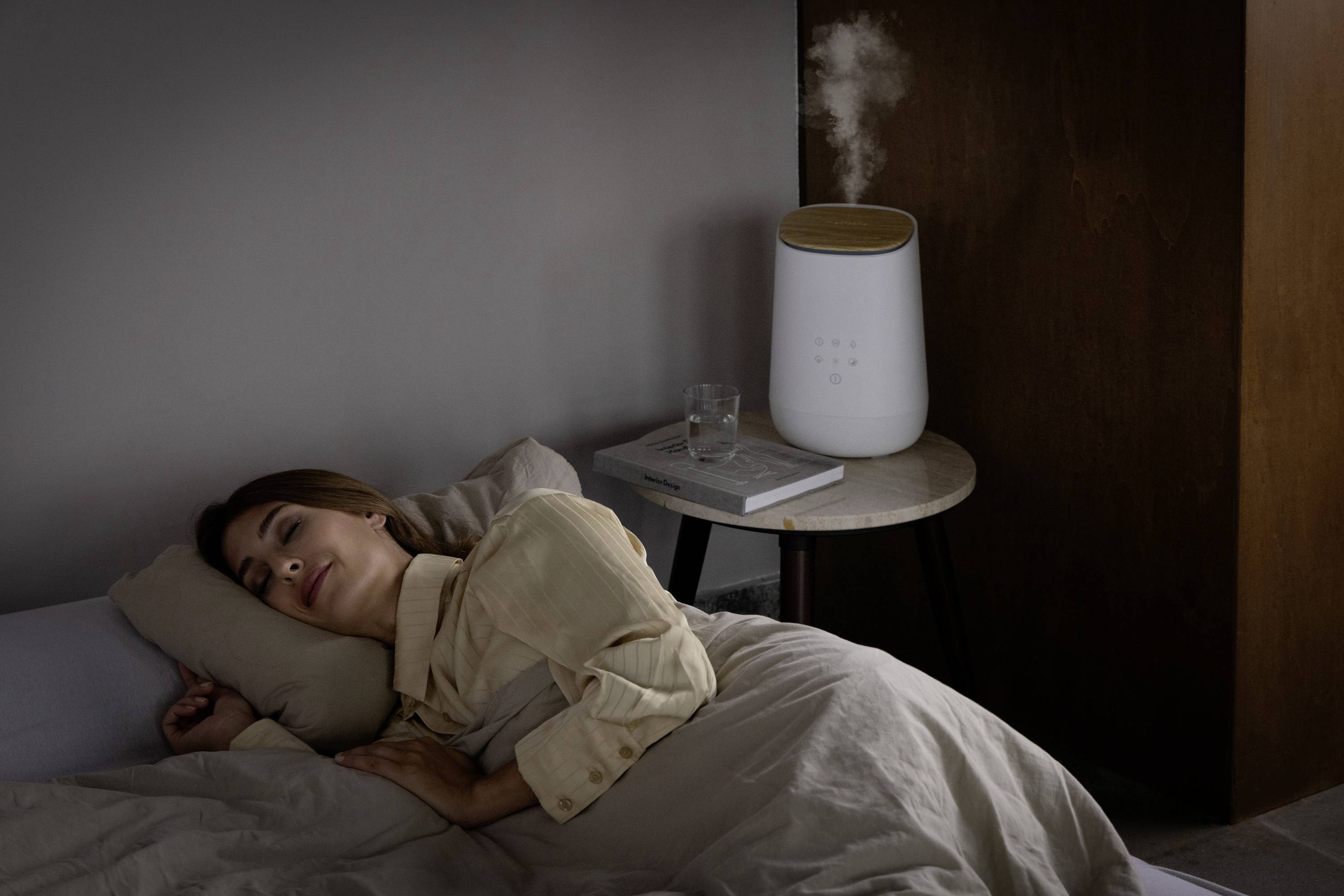 Een vrouw slaapt rustig op een bed, terwijl een luchtbevochtiger op een nachtkastje in de buurt werkt.