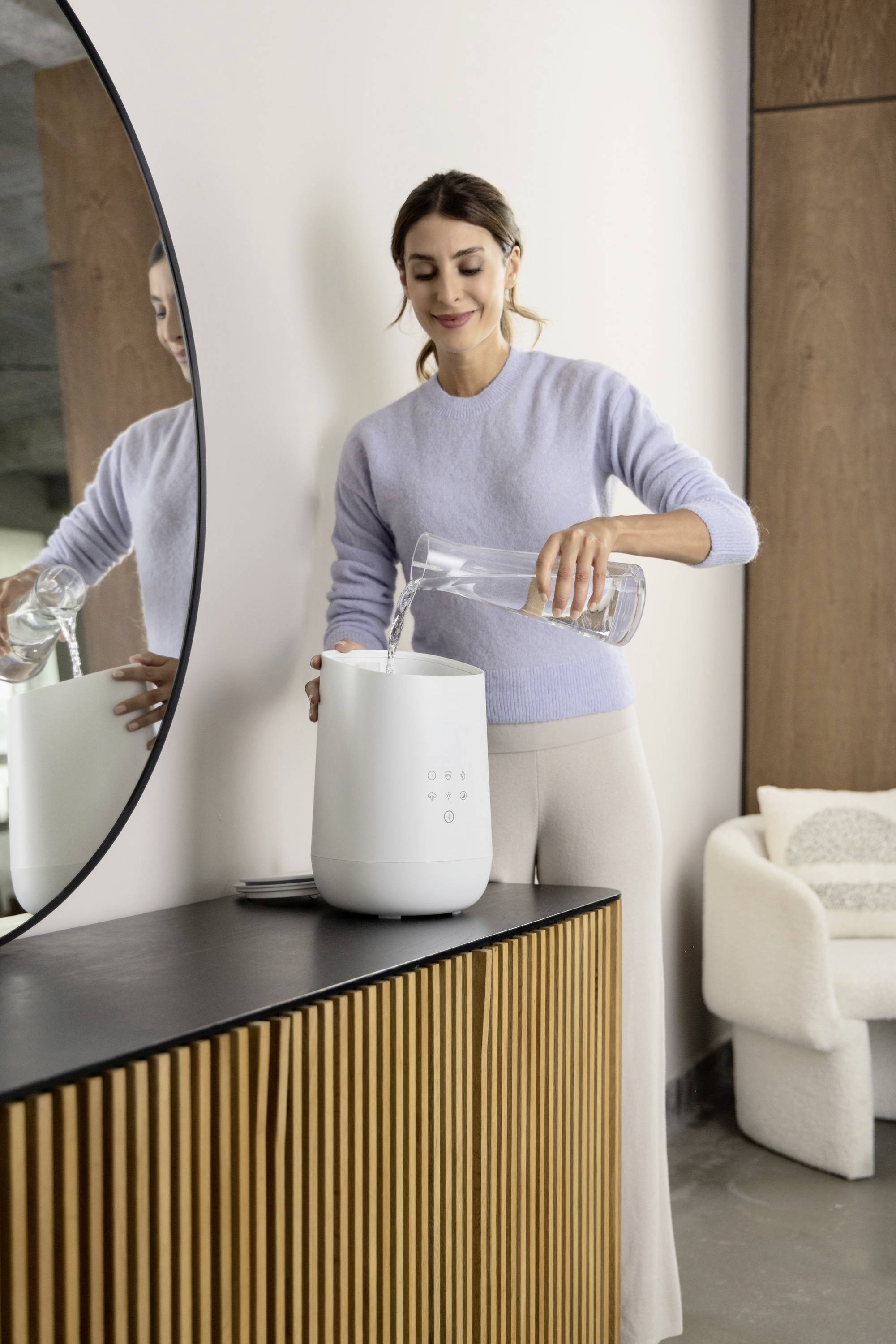 Vrouw in grijze trui giet water uit een kan in een luchtbevochtiger op een modern, elegant dressoir.