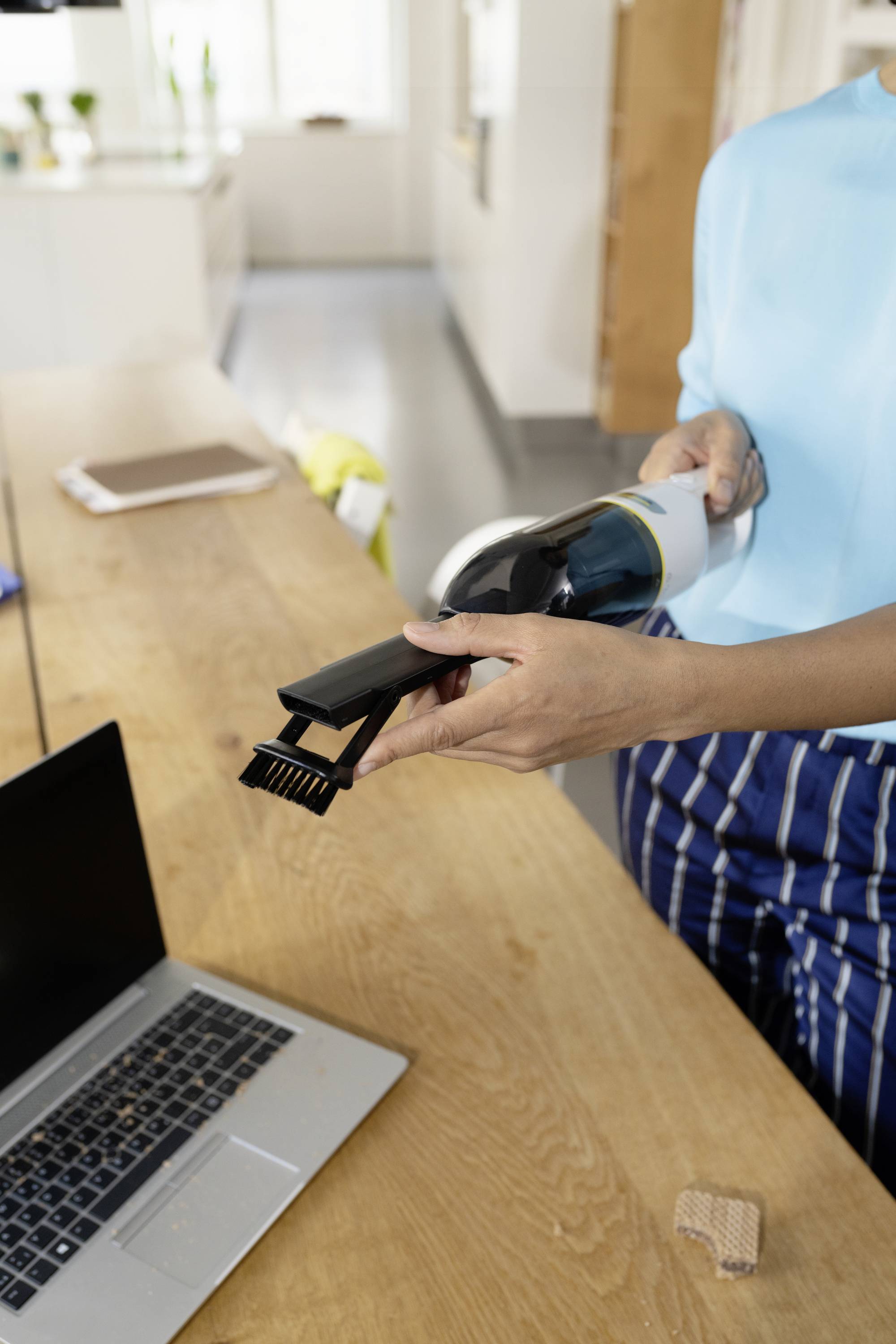 Een persoon houdt een handstofzuiger boven een houten tafel naast een laptop, klaar om kruimels op te zuigen.