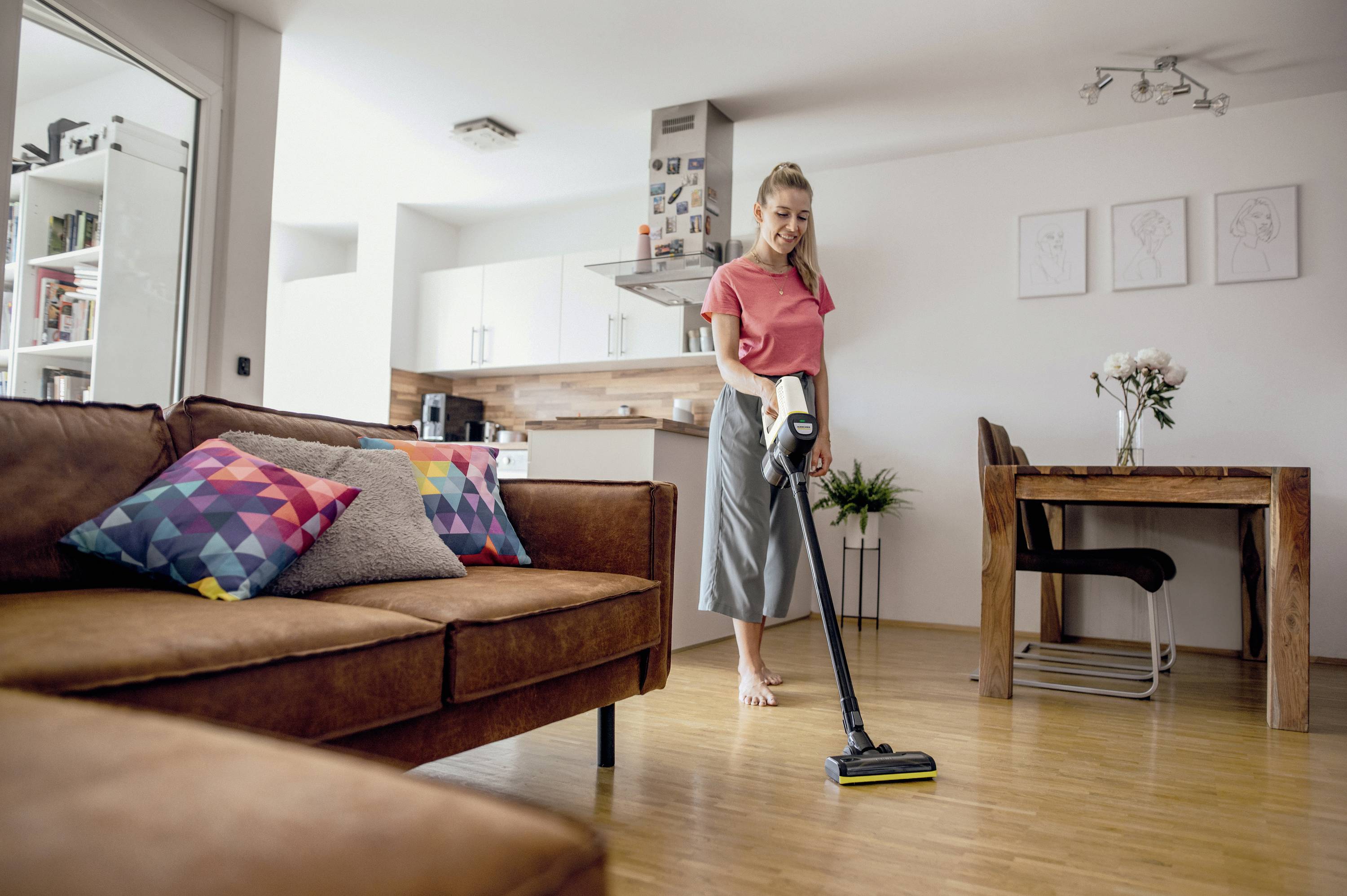 Vrouw stofzuigt de houten vloer in een modern, licht woonvertrek. Op de achtergrond zijn een houten tafel, stoelen en een keuken zichtbaar.