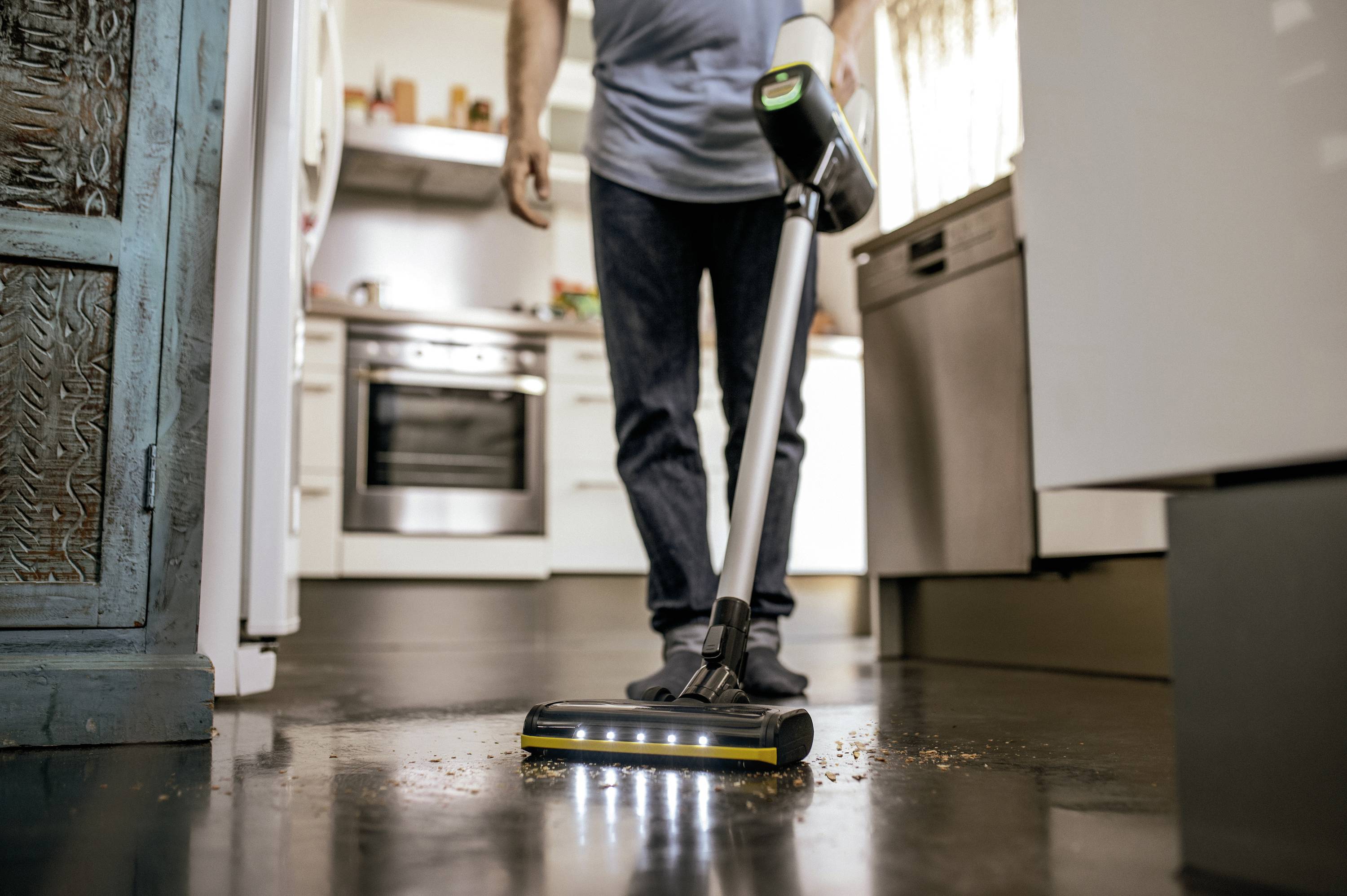 Een persoon reinigt een donkere keukenvloer met een stofzuiger-achtig apparaat. Keuken op de achtergrond; focus op het schoonmaken.